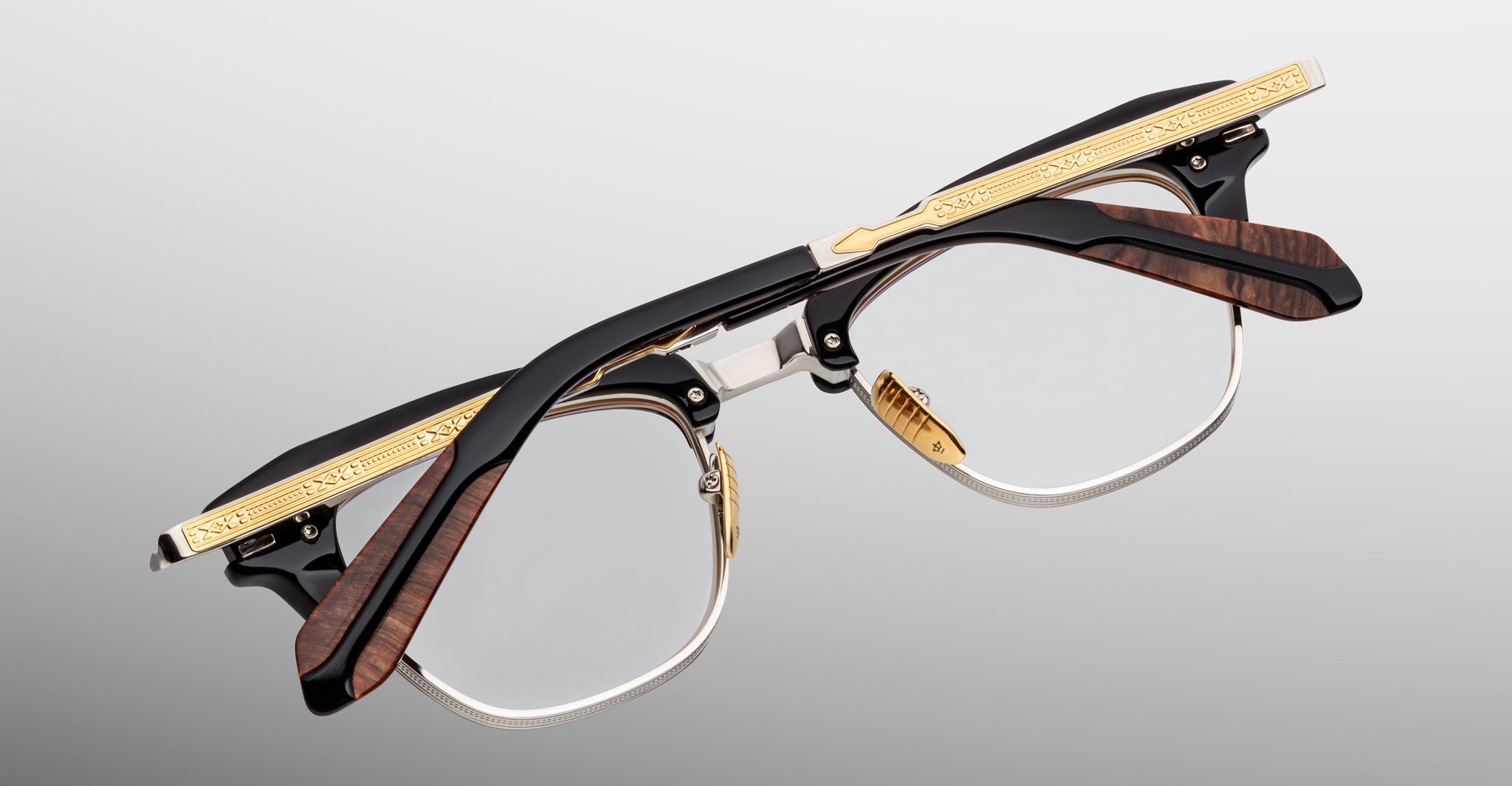 A pair of Jacques Marie Mage Sartre glasses featuring black, gold, and brown wood-textured clubmaster frames is shown upside down against a gray gradient background.