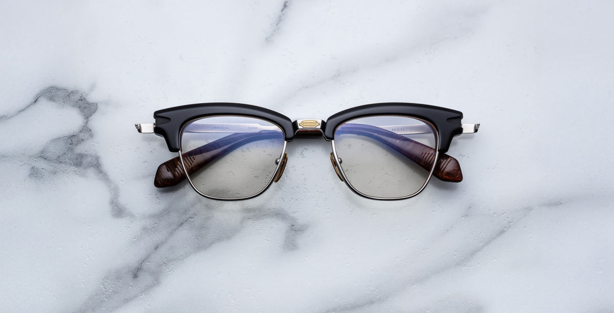 The Jacques Marie Mage Sartre glasses by Jacques Marie Mage, featuring a black and silver clubmaster optical frame with clear lenses, rest on a white marble surface.