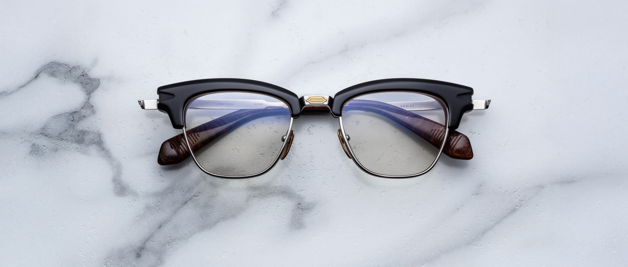 The Jacques Marie Mage Sartre glasses by Jacques Marie Mage, featuring a black and silver clubmaster optical frame with clear lenses, rest on a white marble surface.