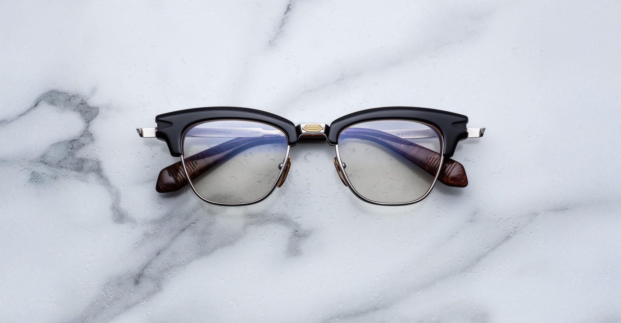 The Jacques Marie Mage Sartre glasses by Jacques Marie Mage, featuring a black and silver clubmaster optical frame with clear lenses, rest on a white marble surface.