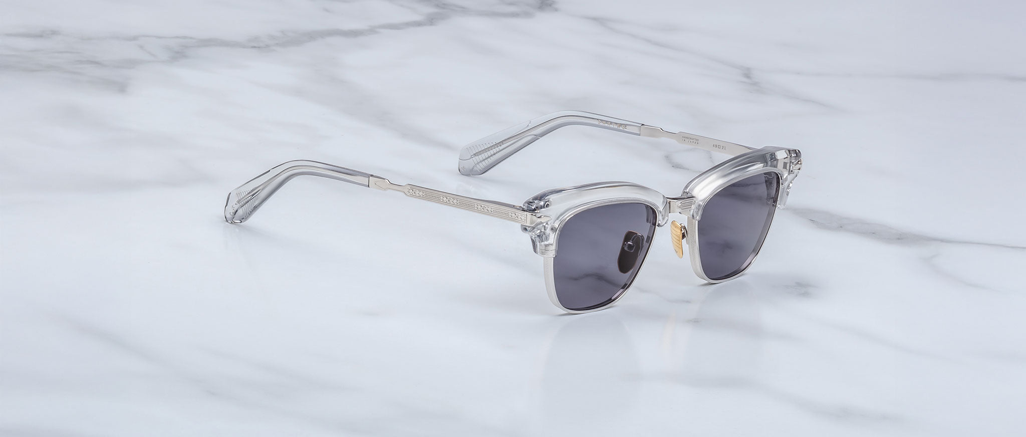 The Jacques Marie Mage Sartre sunglasses by Jacques Marie Mage, featuring clear frames and dark lenses inspired by the iconic clubmaster design, are displayed on a white marble surface.