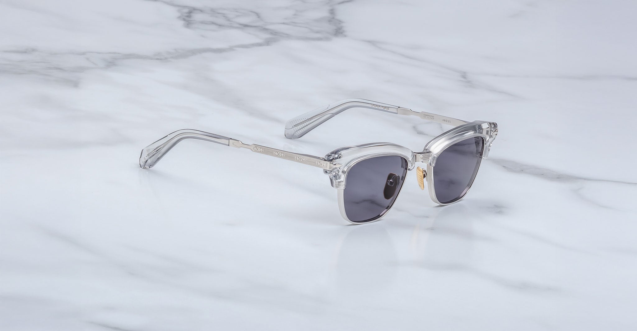 The Jacques Marie Mage Sartre sunglasses by Jacques Marie Mage, featuring clear frames and dark lenses inspired by the iconic clubmaster design, are displayed on a white marble surface.