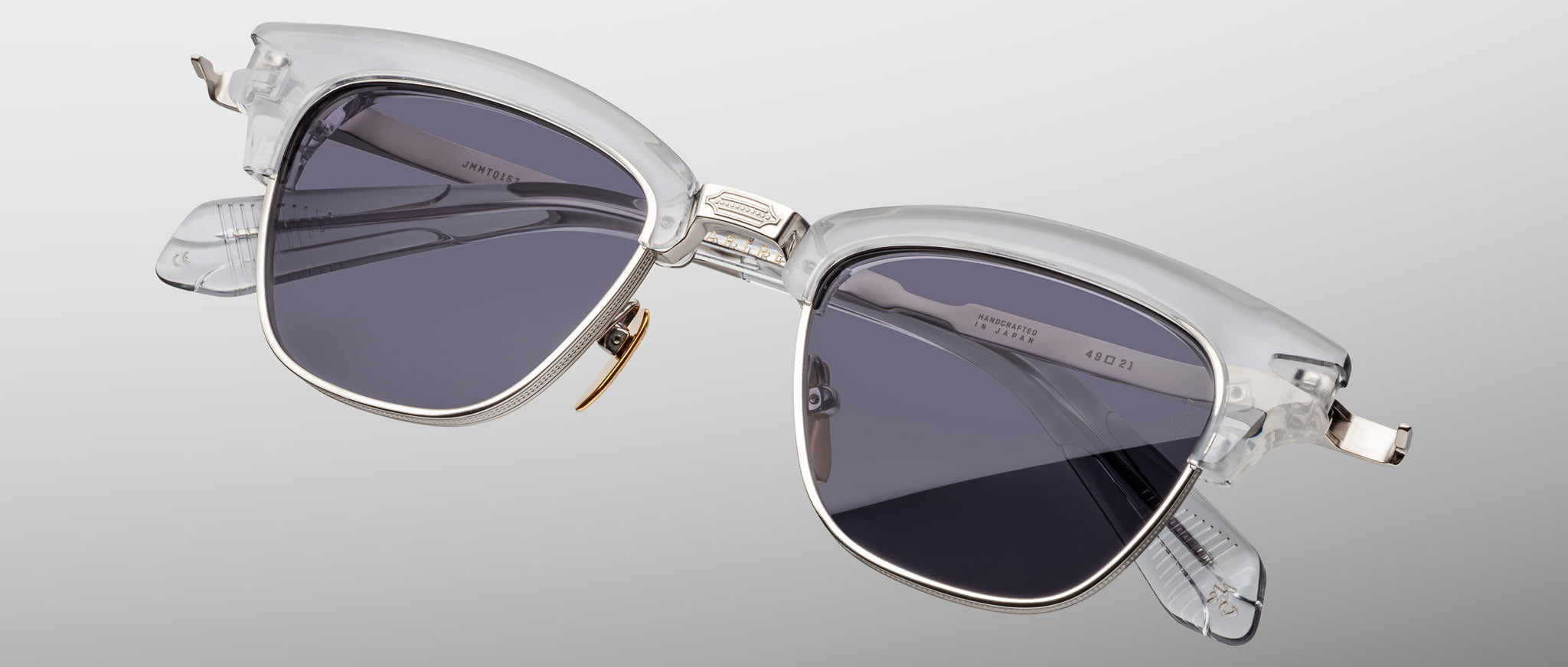 Jacques Marie Mage Sartre sunglasses by Jacques Marie Mage feature clear frames, dark lenses, and metal accents, shown against a plain light gray background.