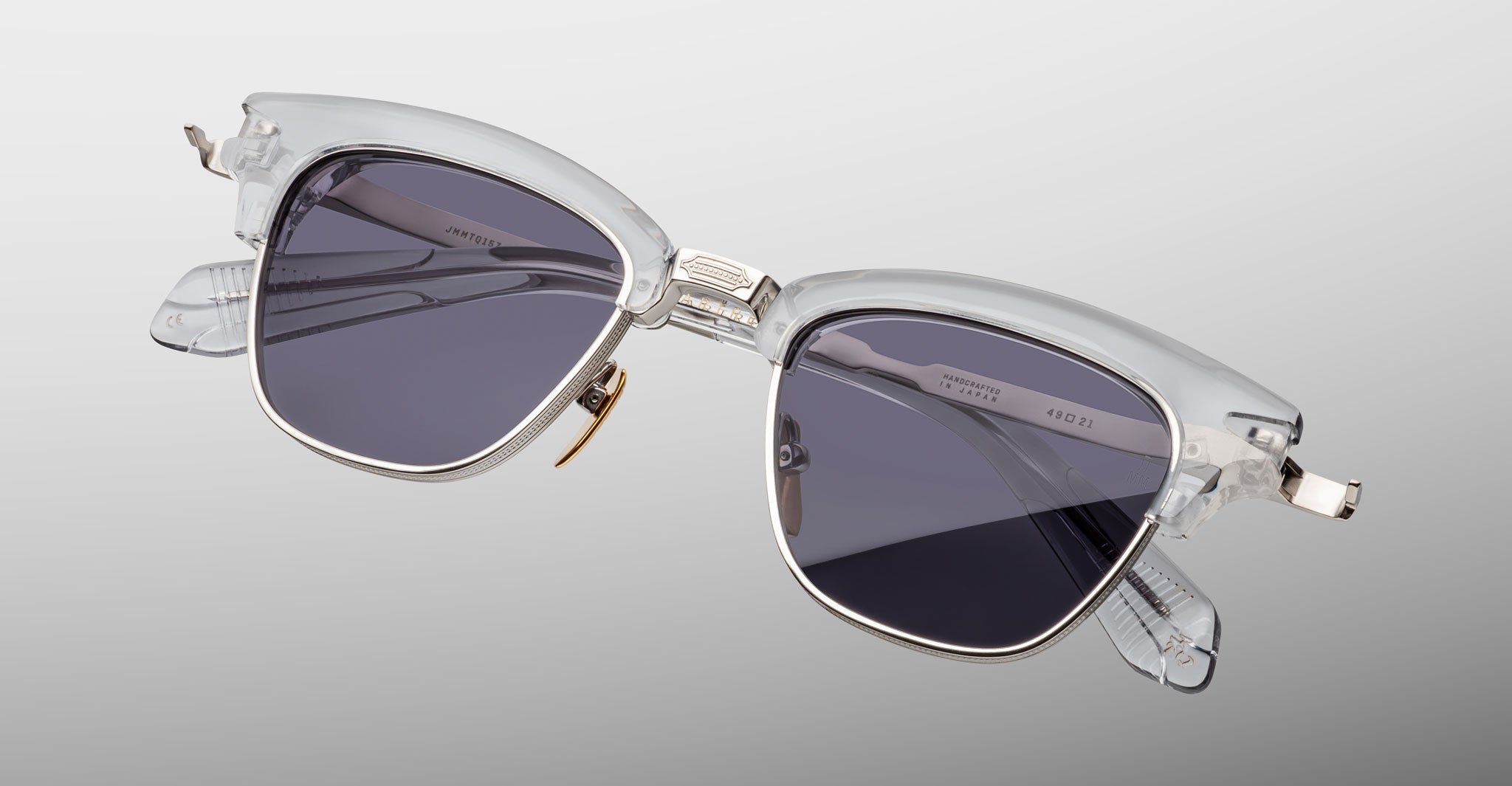 Jacques Marie Mage Sartre sunglasses by Jacques Marie Mage feature clear frames, dark lenses, and metal accents, shown against a plain light gray background.