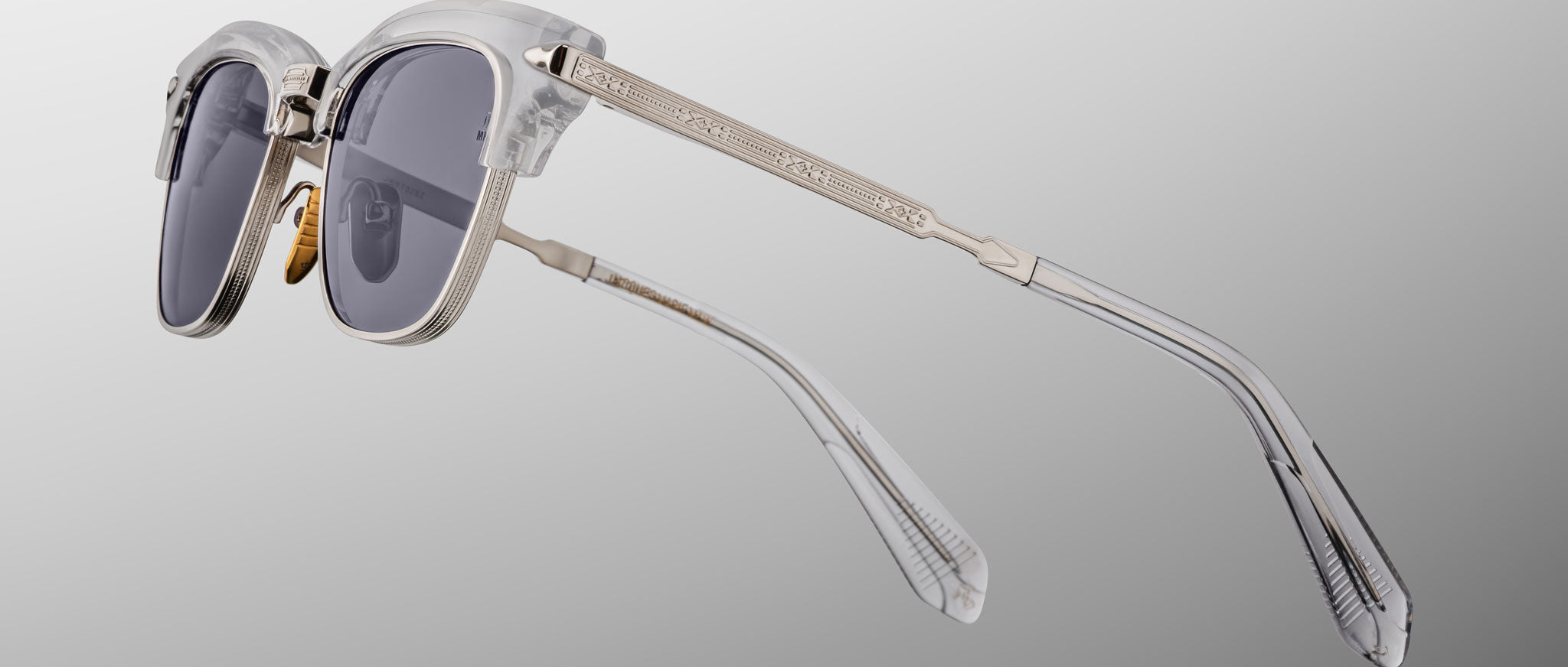 Jacques Marie Mage Sartre sunglasses feature clear frames, dark lenses, and gold-toned metal arms, capturing classic style by Jacques Marie Mage, displayed on a light gray gradient background.