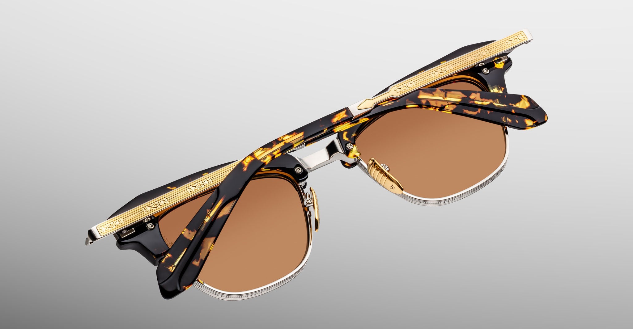 The Jacques Marie Mage Sartre sunglasses by Jacques Marie Mage feature tortoiseshell and gold rims with brown lenses, inspired by classic Sartre glasses, pictured folded on a neutral gray background.