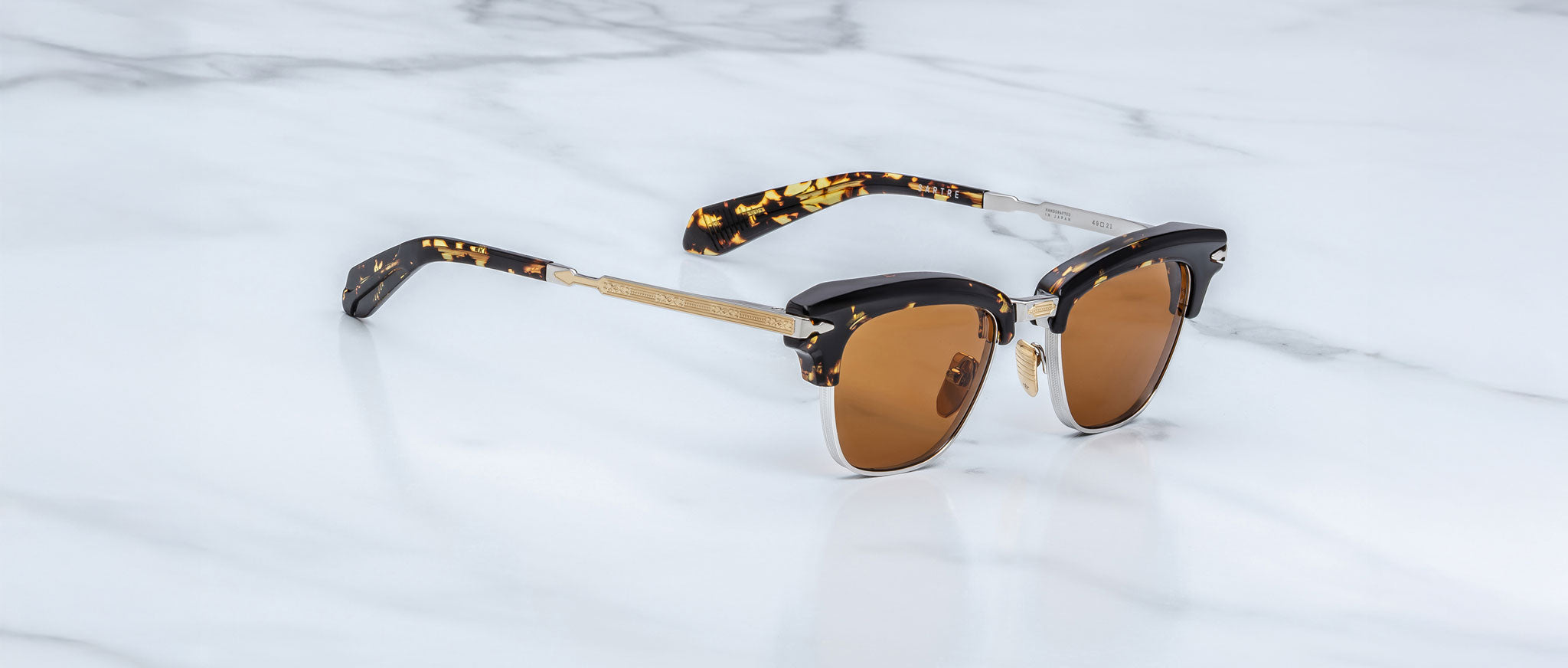 A pair of Jacques Marie Mage Sartre glasses featuring brown-tinted lenses, gold accents, and black tortoiseshell arms is displayed on a white marble surface.