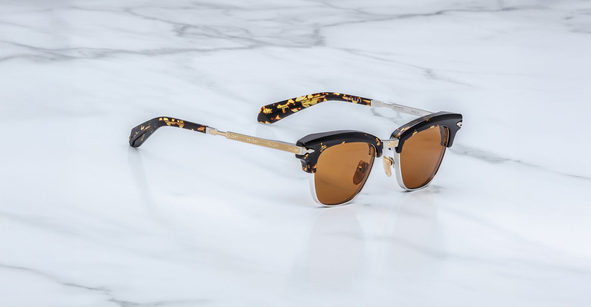 A pair of Jacques Marie Mage Sartre glasses featuring brown-tinted lenses, gold accents, and black tortoiseshell arms is displayed on a white marble surface.
