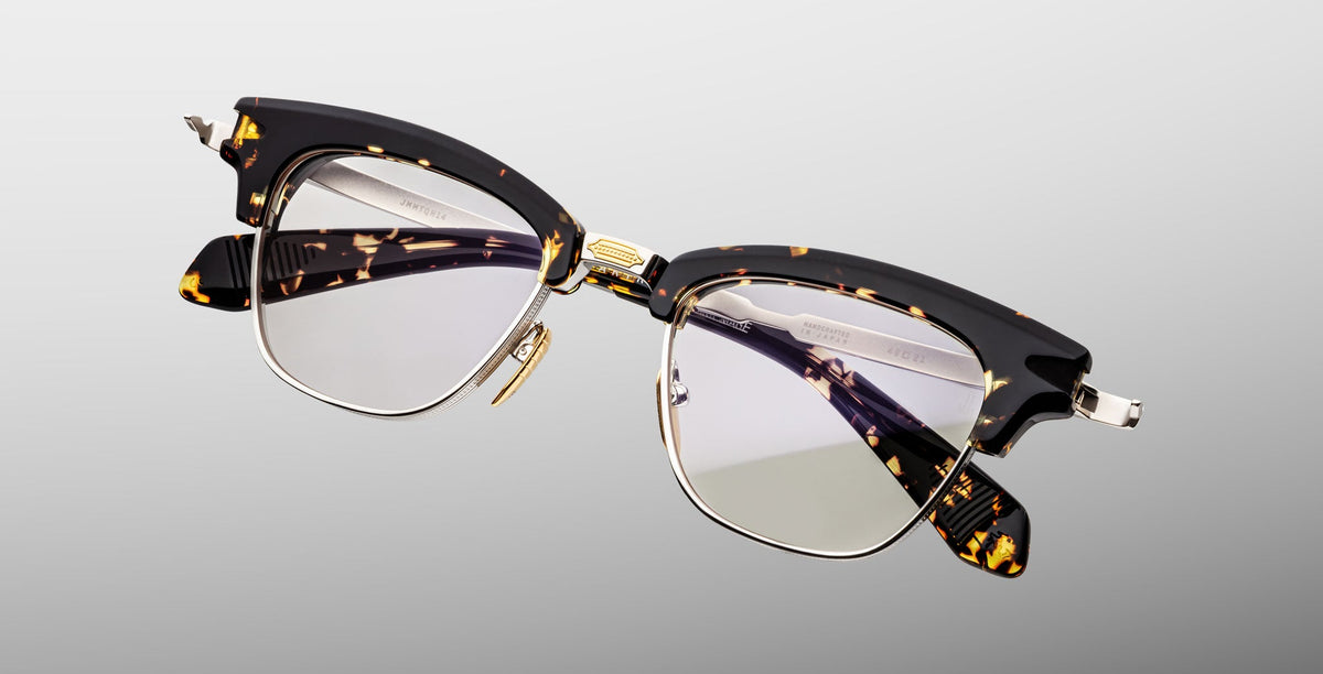 The Jacques Marie Mage Sartre by Jacques Marie Mage are clubmaster eyeglasses in tortoiseshell and black with clear lenses, gold detailing, and metal accents, displayed on a light gray background.