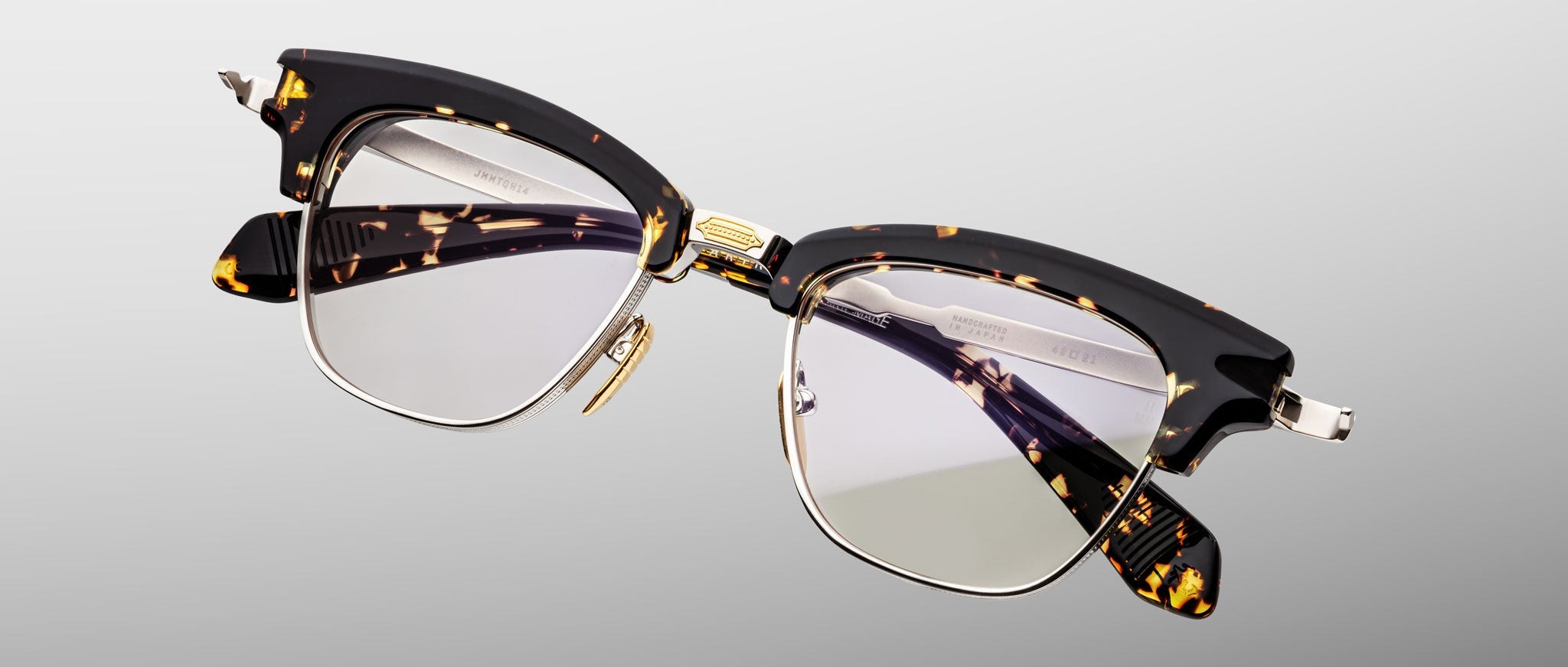 The Jacques Marie Mage Sartre by Jacques Marie Mage are clubmaster eyeglasses in tortoiseshell and black with clear lenses, gold detailing, and metal accents, displayed on a light gray background.