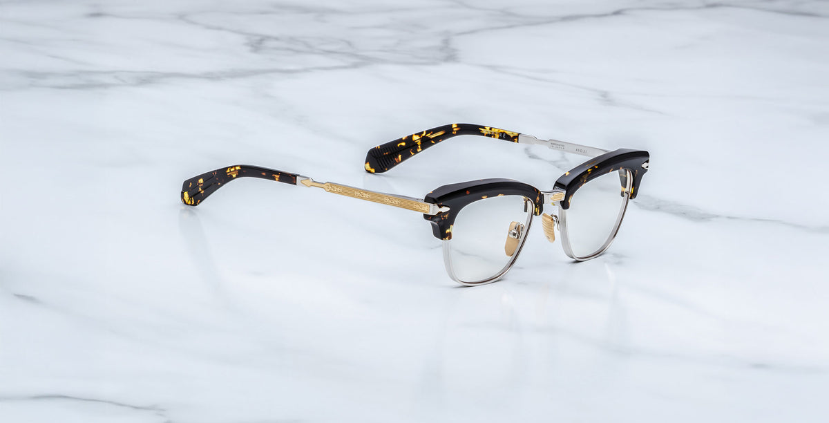Jacques Marie Mage Sartre optical eyeglasses by Jacques Marie Mage, featuring gold metal rims and black tortoiseshell arms, are displayed on a white marble surface.