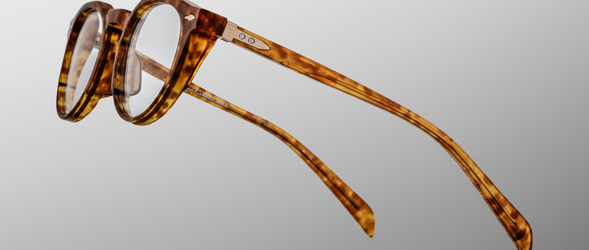 The Jacques Marie Mage Sheridan eyeglasses feature a round tortoiseshell frame, clear lenses, metal hinge accents, and gently curved arms, presented against a plain grey background.
