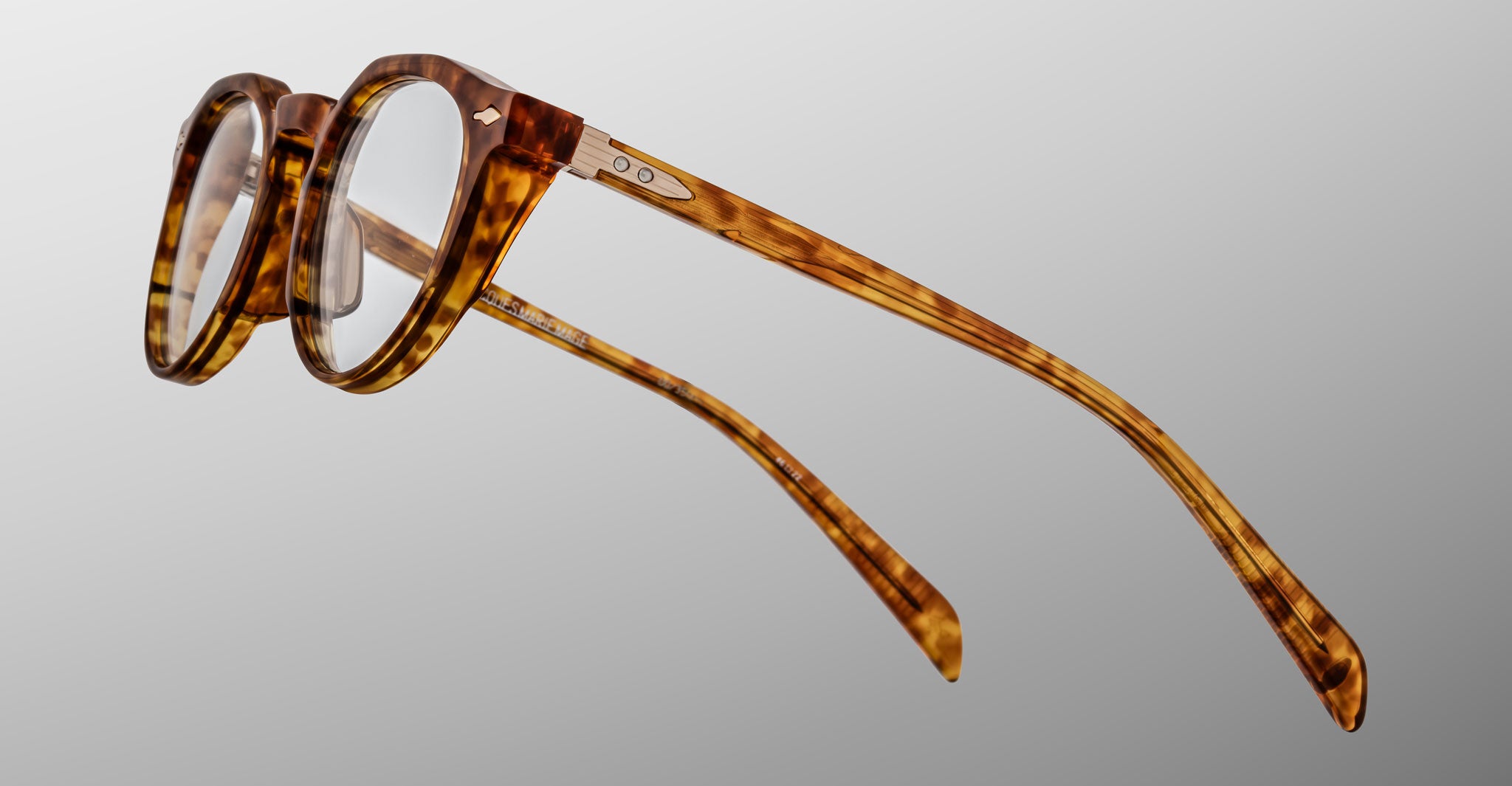 The Jacques Marie Mage Sheridan eyeglasses feature a round tortoiseshell frame, clear lenses, metal hinge accents, and gently curved arms, presented against a plain grey background.
