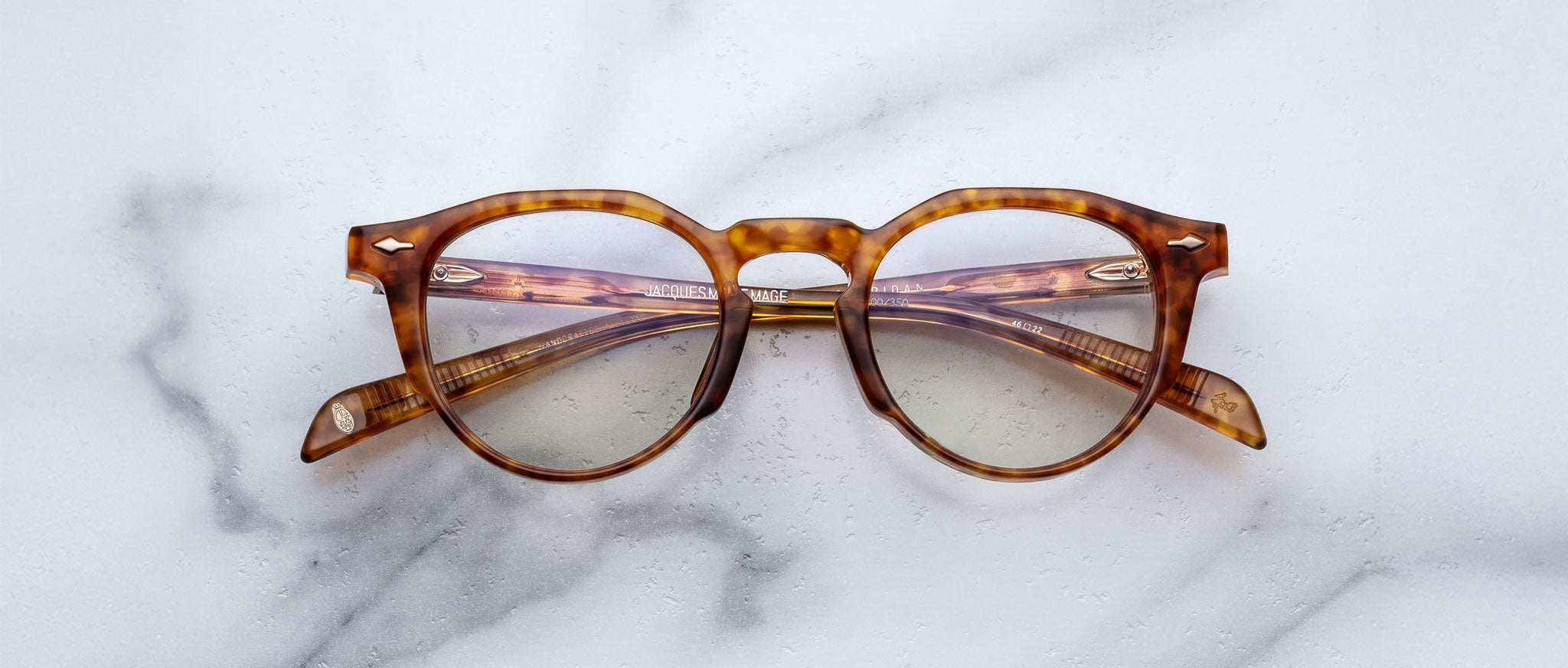 Jacques Marie Mage Sheridan round tortoiseshell eyeglasses with clear lenses displayed on a white marble surface, arms folded outward to reveal frame details.