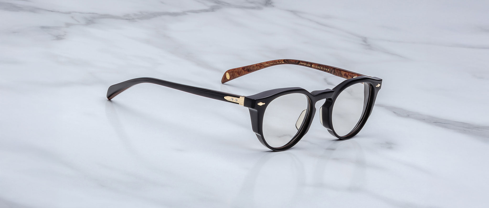 A pair of Jacques Marie Mage Sheridan round black eyeglasses with gold hinge detailing and brown tortoiseshell inner arms rests on a white marble surface.
