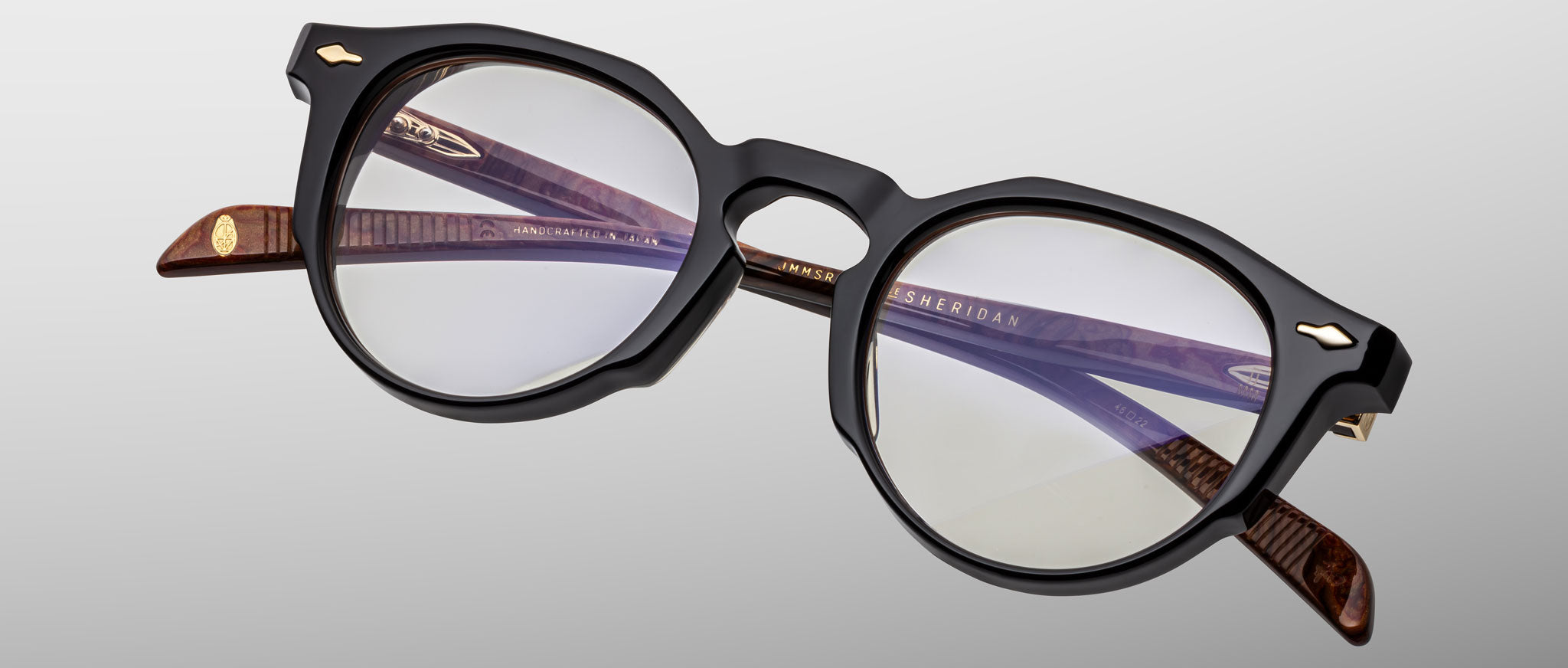 The Jacques Marie Mage Sheridan features round eyeglasses with thick black frames, brown textured temple arms, and clear lenses. Logo details appear on the inner arms, all set against a light gray background.