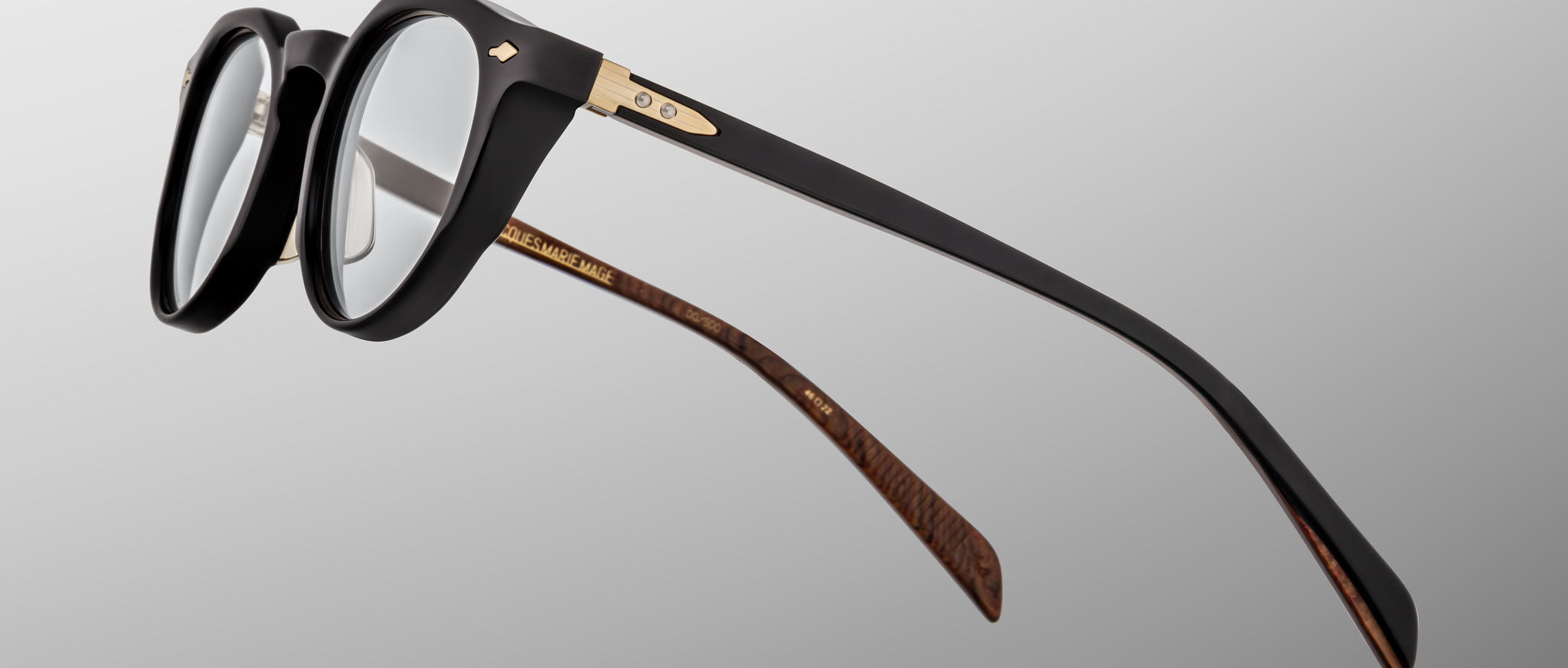 The Jacques Marie Mage Sheridan eyeglasses are shown in close-up, featuring black frames with gold accents and brown patterned inner temples, set against a gray gradient background.