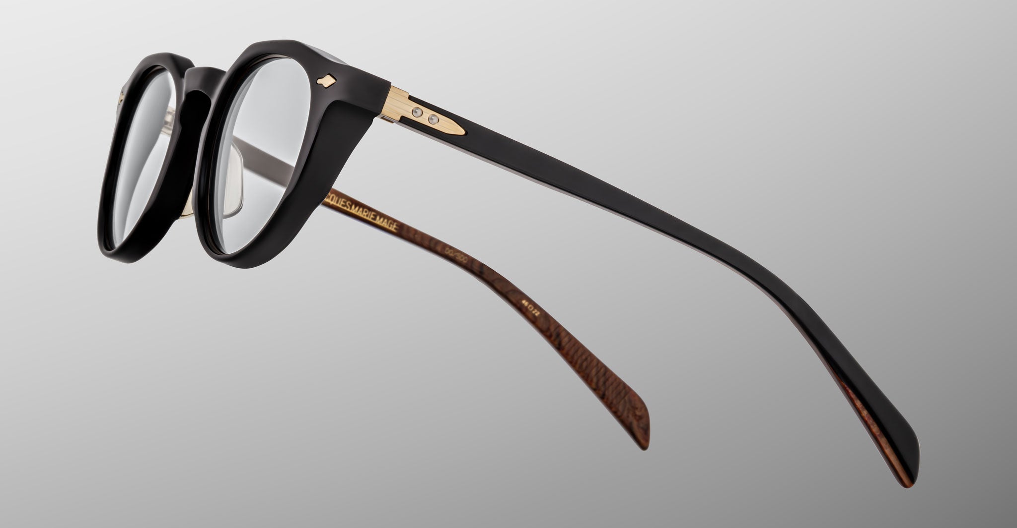 The Jacques Marie Mage Sheridan eyeglasses are shown in close-up, featuring black frames with gold accents and brown patterned inner temples, set against a gray gradient background.