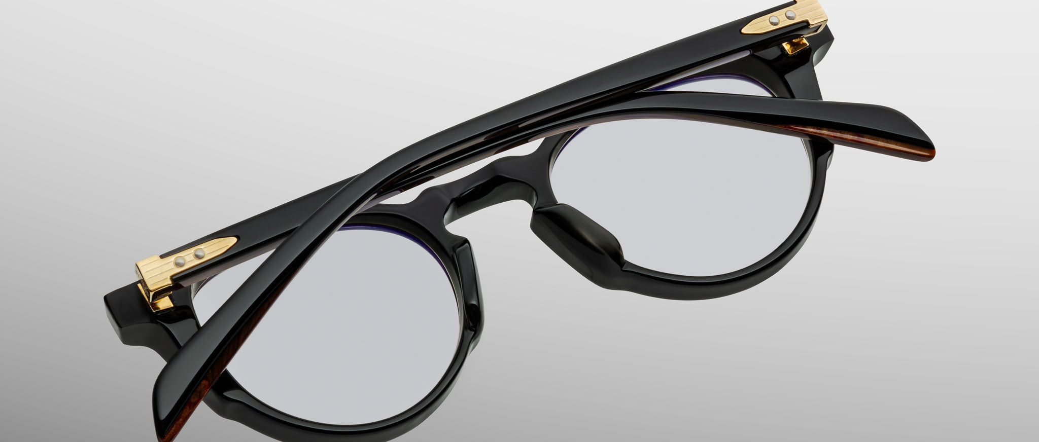 The Jacques Marie Mage Sheridan eyeglasses, featuring a black round frame with gold accents, are shown folded and viewed from above on a light gray background.