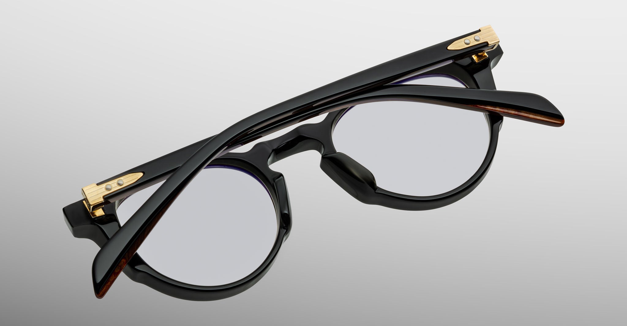 The Jacques Marie Mage Sheridan eyeglasses, featuring a black round frame with gold accents, are shown folded and viewed from above on a light gray background.