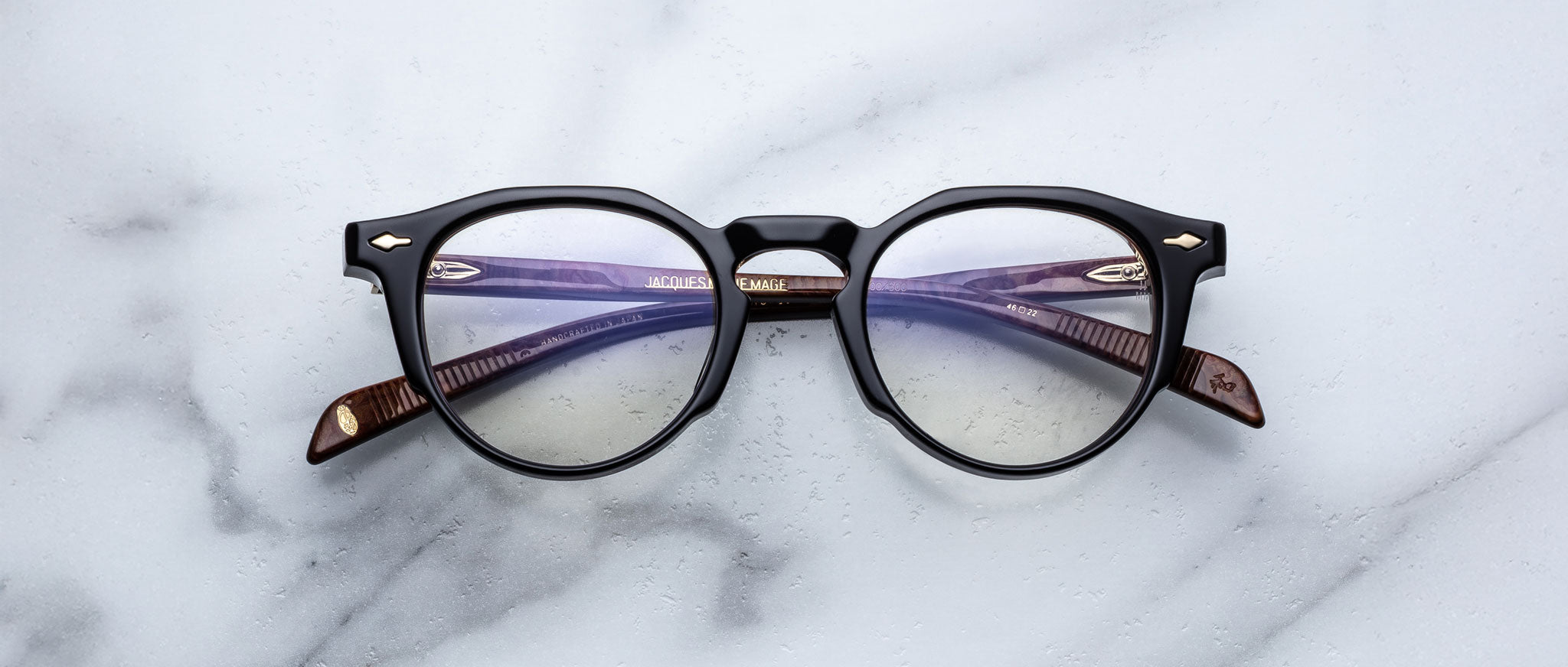 A pair of Jacques Marie Mage Sheridan round black eyeglasses with brown arms is placed on a white marble surface.