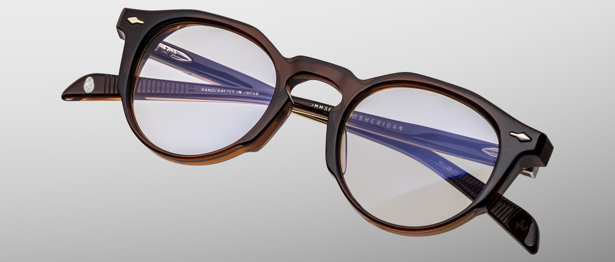 The Jacques Marie Mage Sheridan features round brown acetate frames with clear lenses, blue-tinted inner arms, and silver arrow accents by the hinges, displayed against a light gray background.