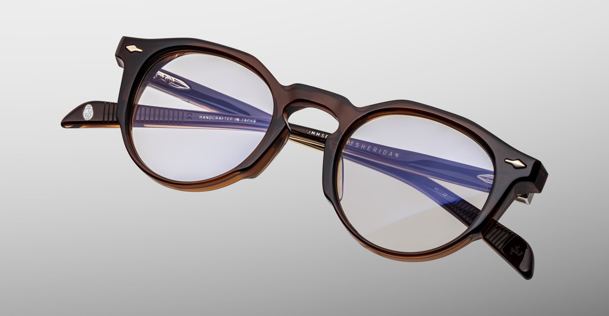 The Jacques Marie Mage Sheridan features round brown acetate frames with clear lenses, blue-tinted inner arms, and silver arrow accents by the hinges, displayed against a light gray background.