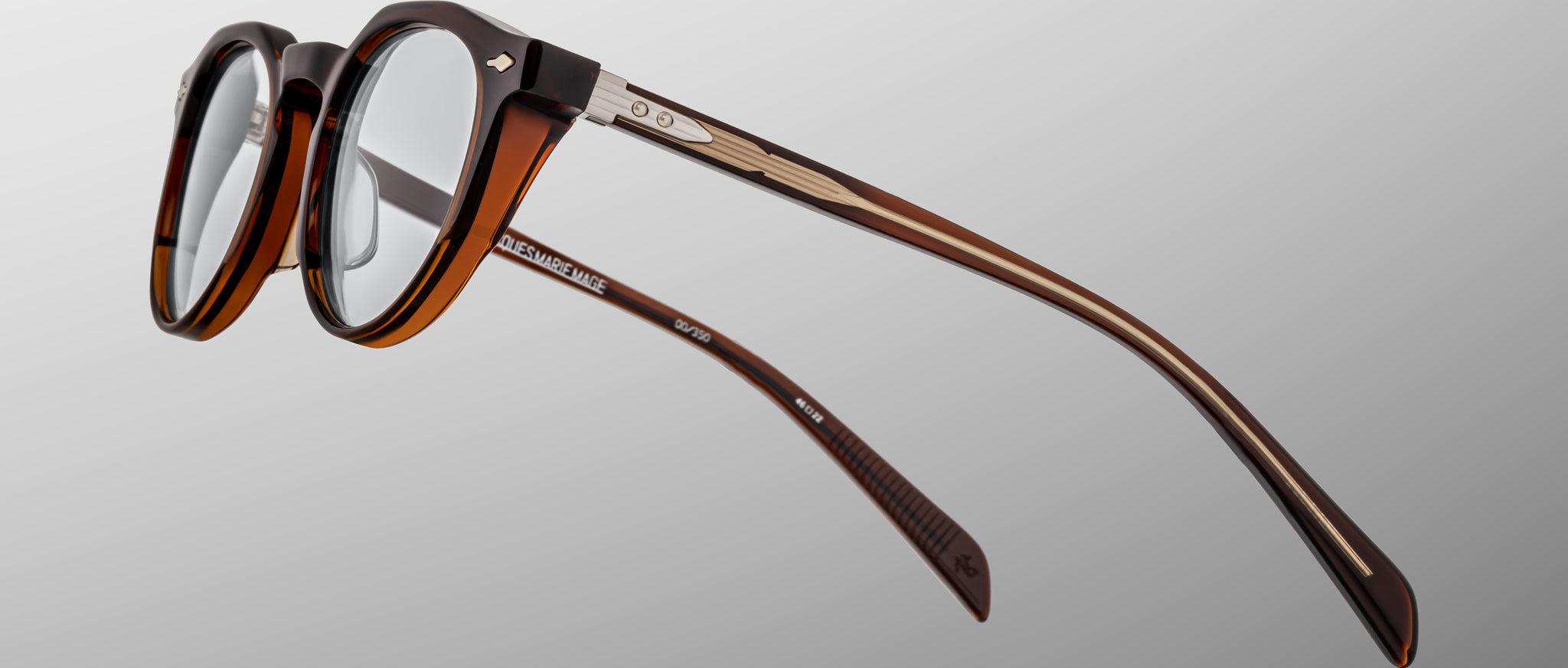 Jacques Marie Mage Sheridan: brown, semi-transparent round eyeglasses with metal temple accents, shown at an angle on a plain gray background.