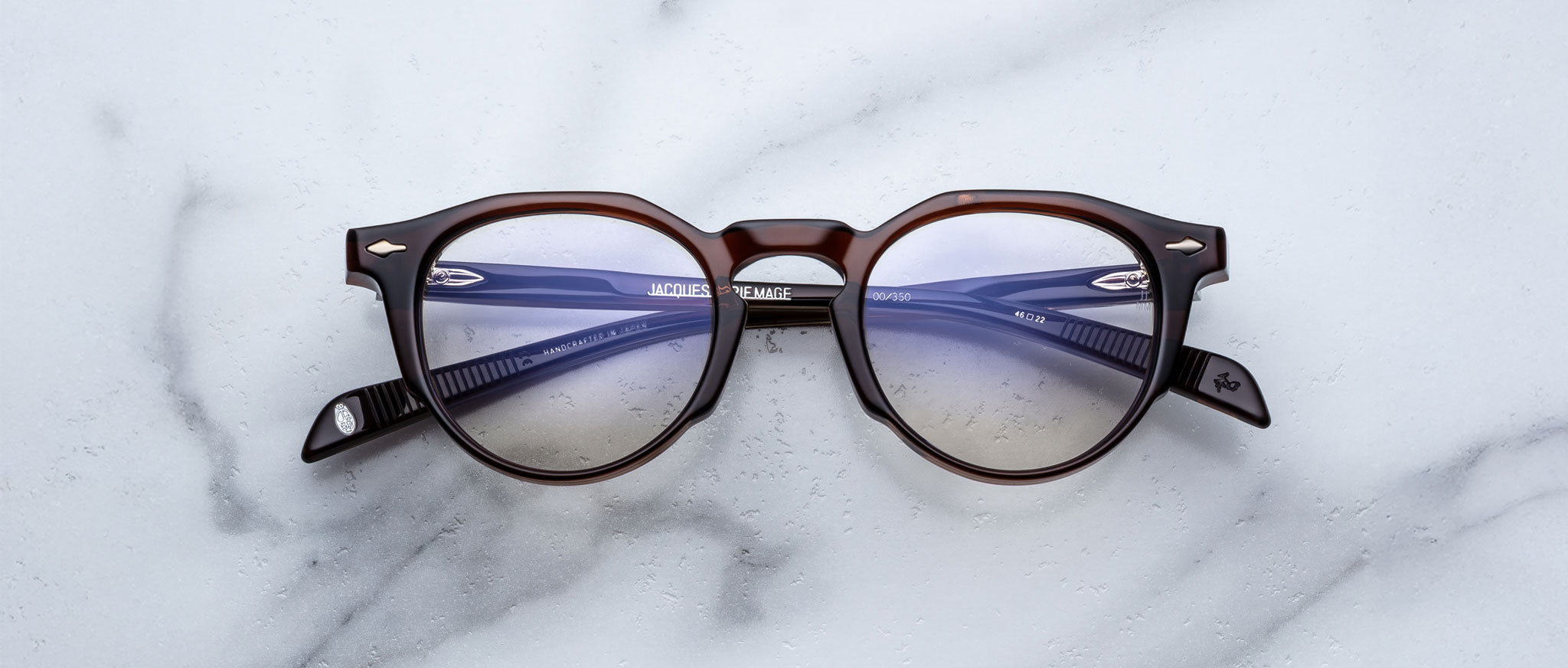 The Jacques Marie Mage Sheridan eyeglasses, featuring round dark brown frames with blue lenses and folded arms, rest on a white marble surface.