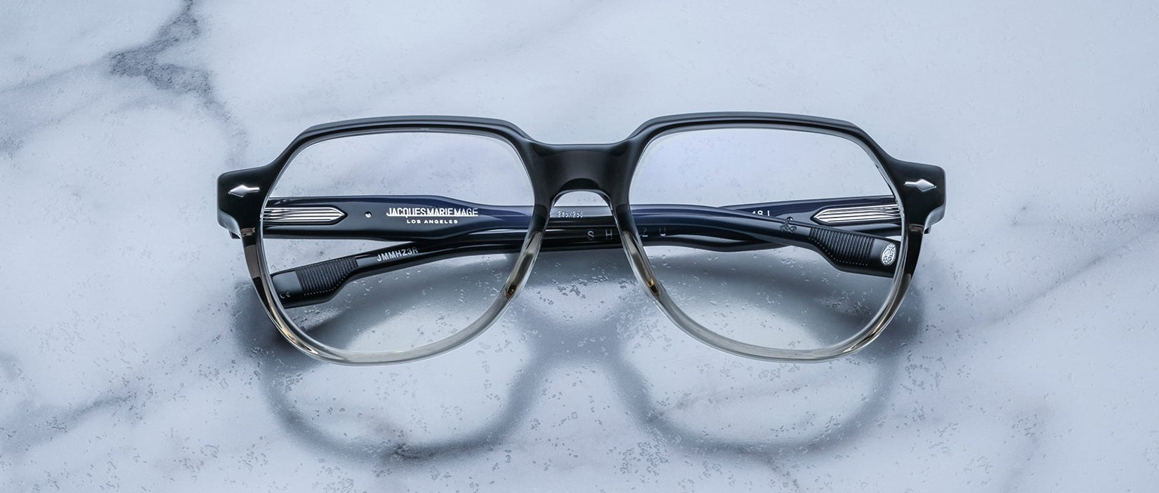 A pair of Jacques Marie Mage Shozo black and clear rectangular eyeglasses are folded and placed on a light gray marble surface.