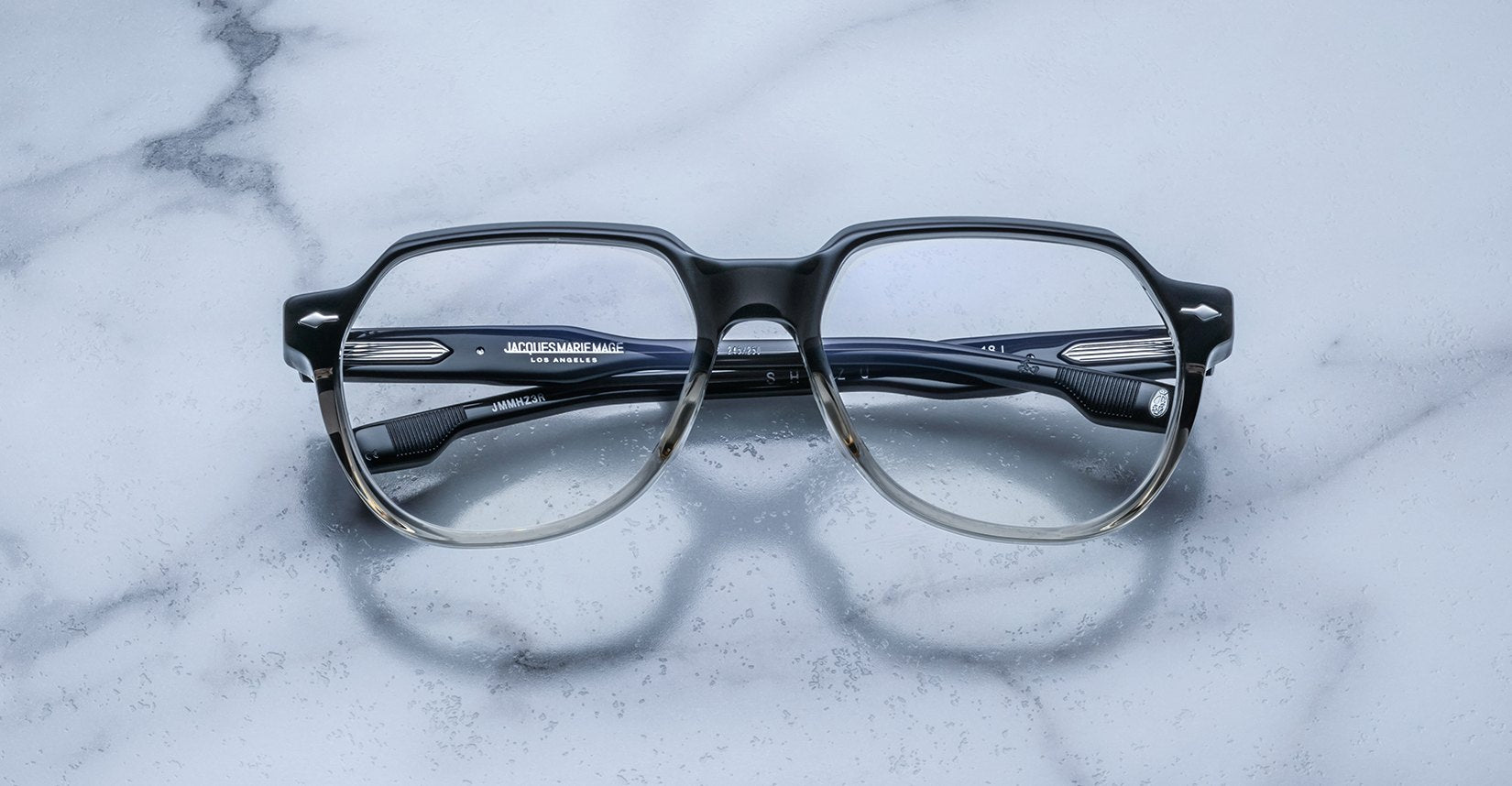 A pair of Jacques Marie Mage Shozo black and clear rectangular eyeglasses are folded and placed on a light gray marble surface.