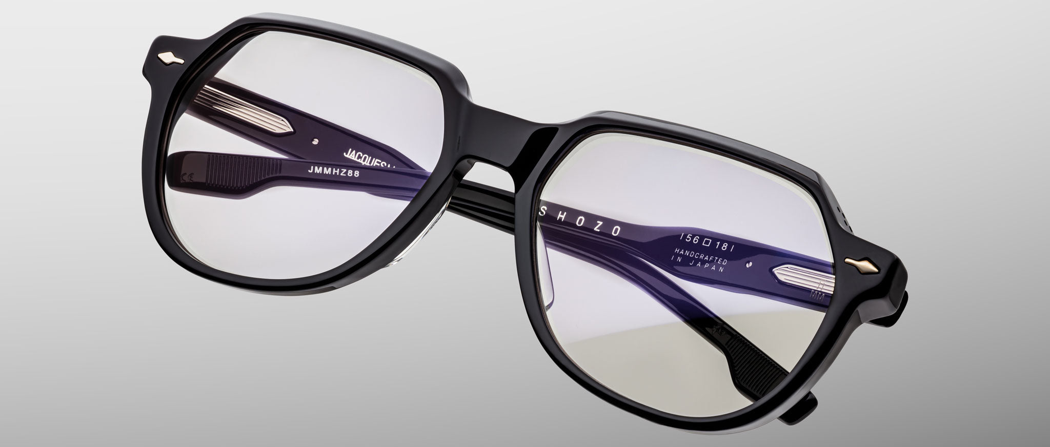 The Jacques Marie Mage Shozo features black-rimmed eyeglasses with clear lenses and a modern geometric frame, displayed against a light gray gradient background. Branding text appears on the inside of the temples.