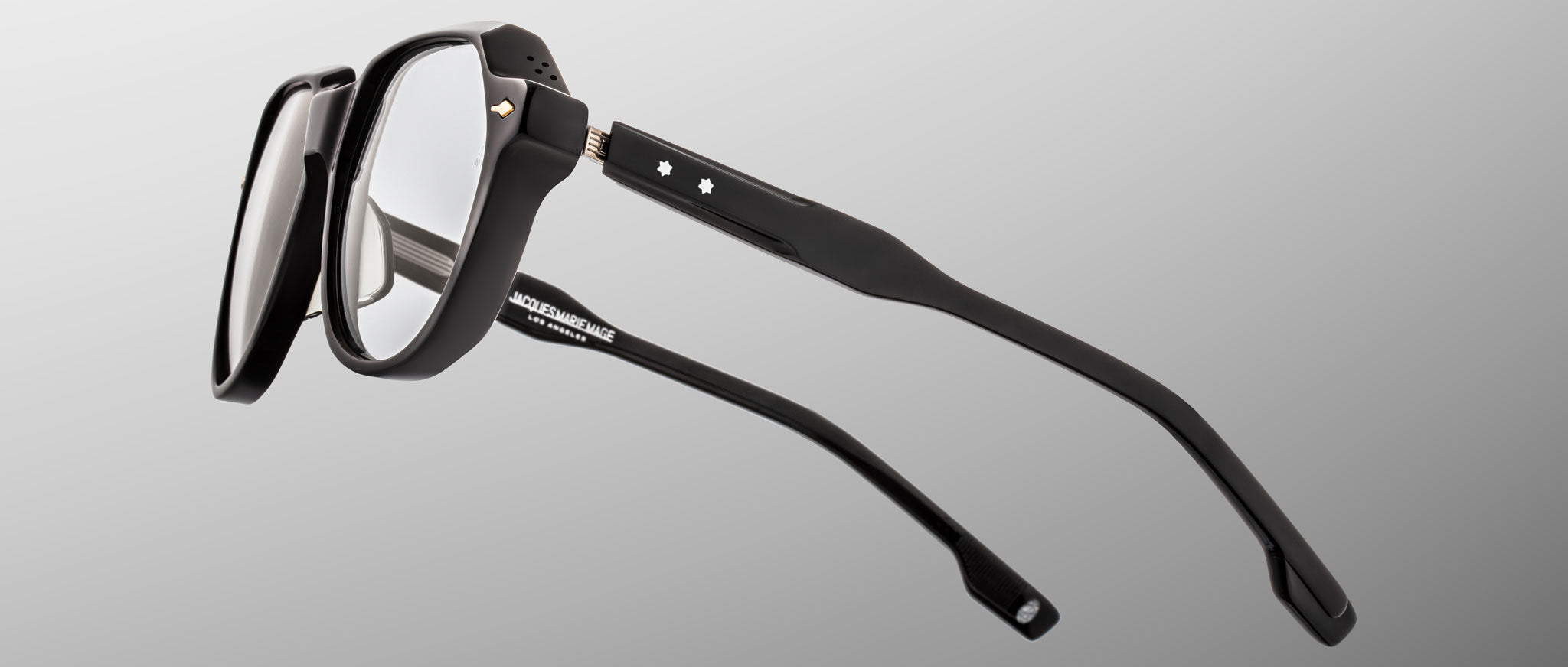 The Jacques Marie Mage Shozo eyeglasses feature black thick-rimmed frames with clear lenses, viewed from an angled side perspective on a light gray background. Two small white star accents adorn the temples.