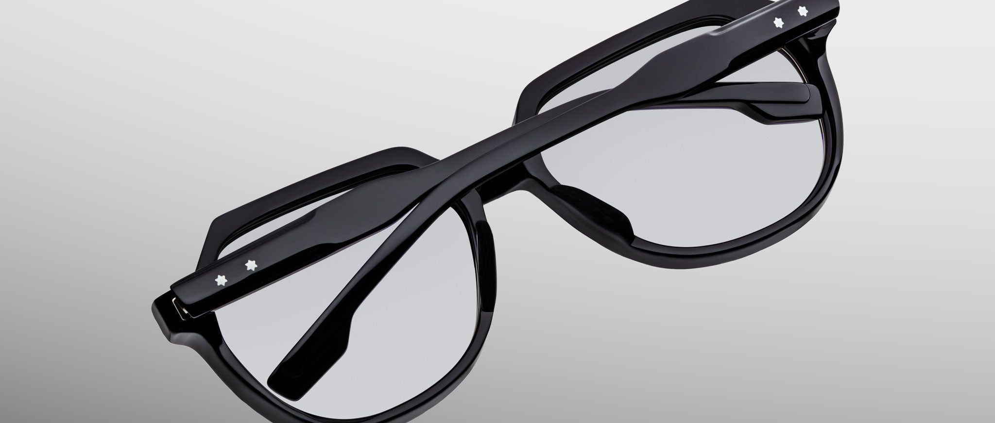 The Jacques Marie Mage Shozo glasses, black with thick rims and folded arms, feature small white star-shaped accents on the arms and are shown from above against a light gray gradient background.