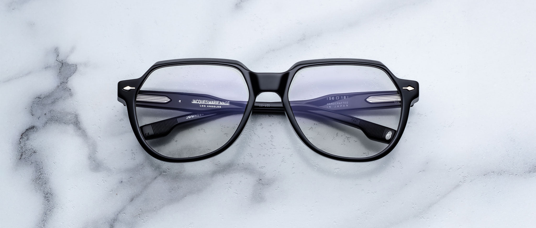 The Jacques Marie Mage Shozo eyeglasses, featuring black frames and clear lenses, rest on a white marble surface. The temples display brand and size details, and the frames have a subtle shine.