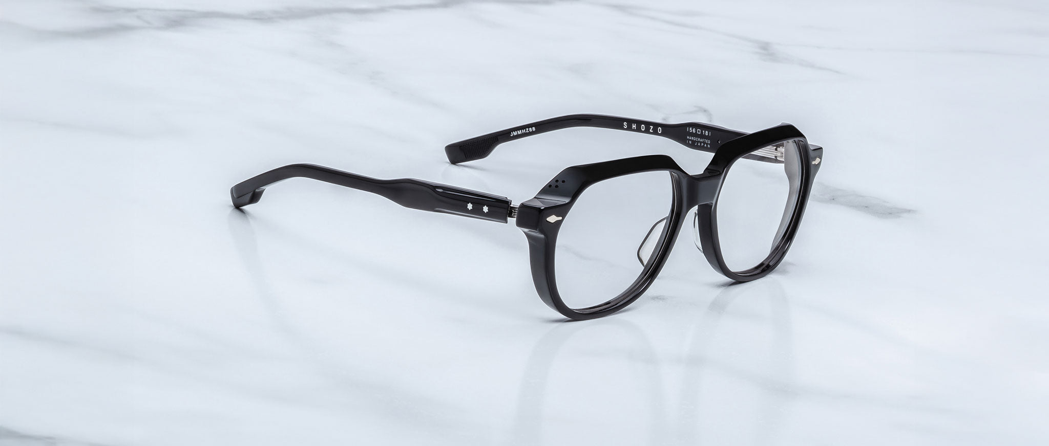 Jacques Marie Mage Shozo eyeglasses with clear lenses and thick black frames rest on a white marble surface. The arms are slightly bent, featuring metallic accents near the hinges.