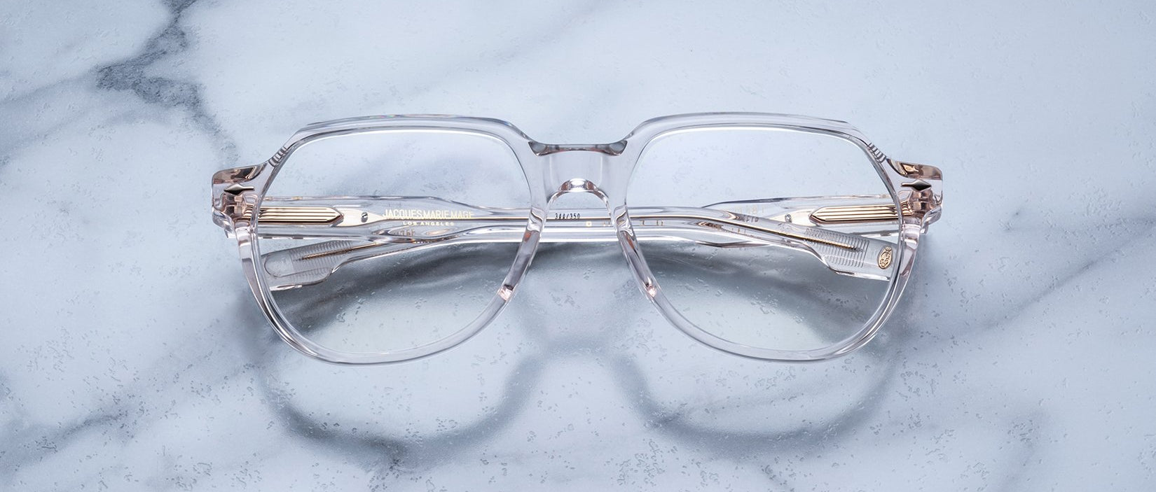 Jacques Marie Mage Shozo clear-framed eyeglasses with gold accents are displayed on a light gray marble surface, seen from above.