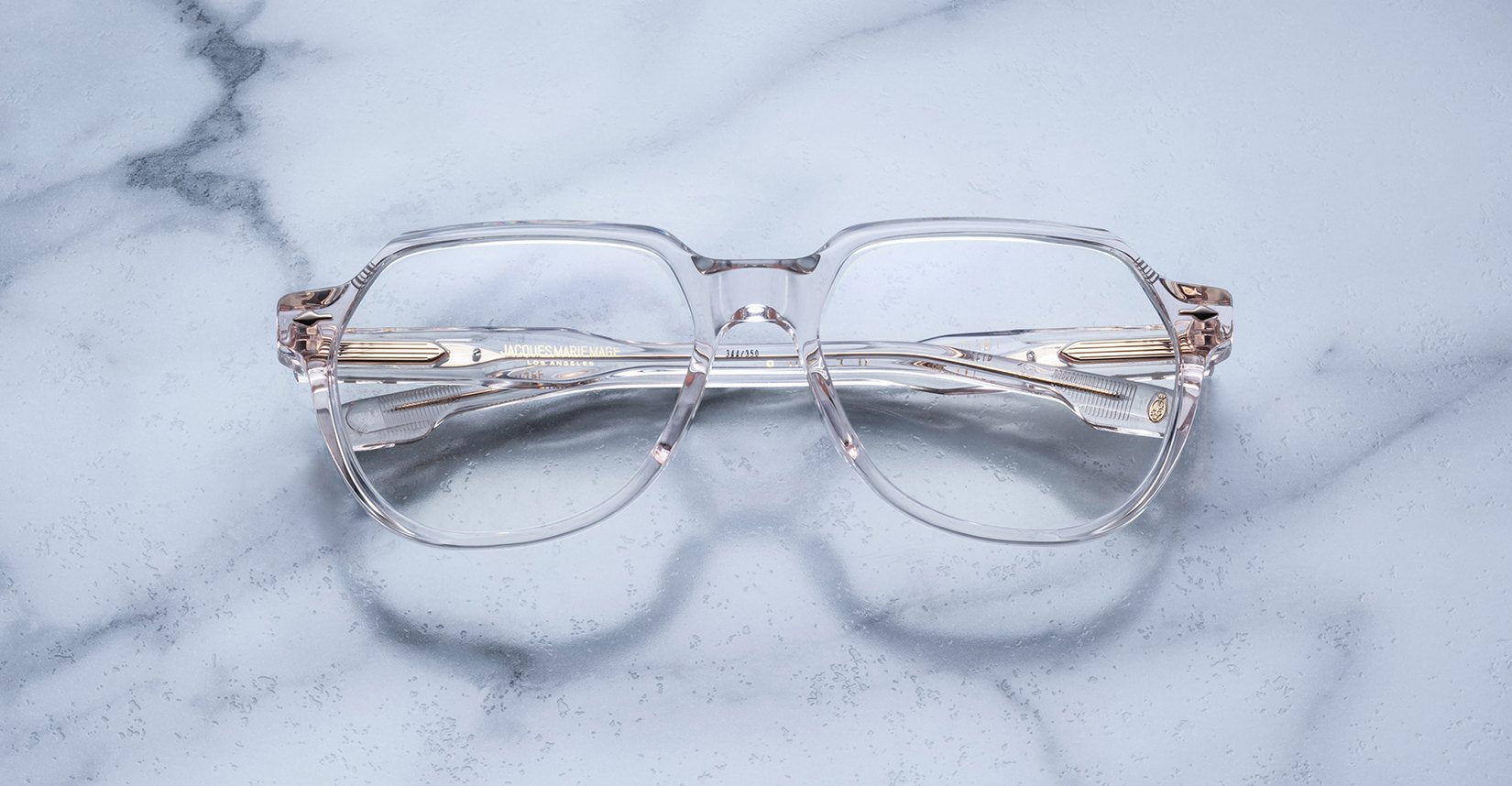 Jacques Marie Mage Shozo clear-framed eyeglasses with gold accents are displayed on a light gray marble surface, seen from above.