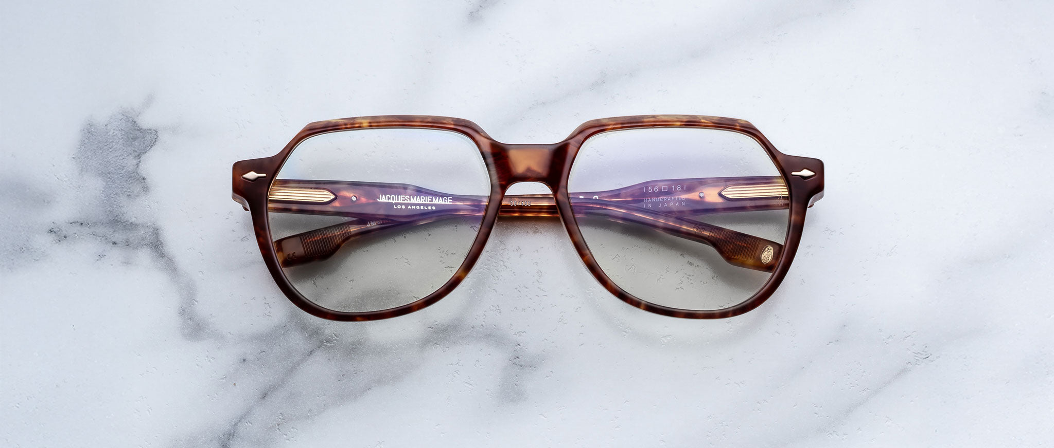 The Jacques Marie Mage Shozo eyeglasses, featuring a brown geometric frame and purple-tinted arms, rest on a white marble surface.
