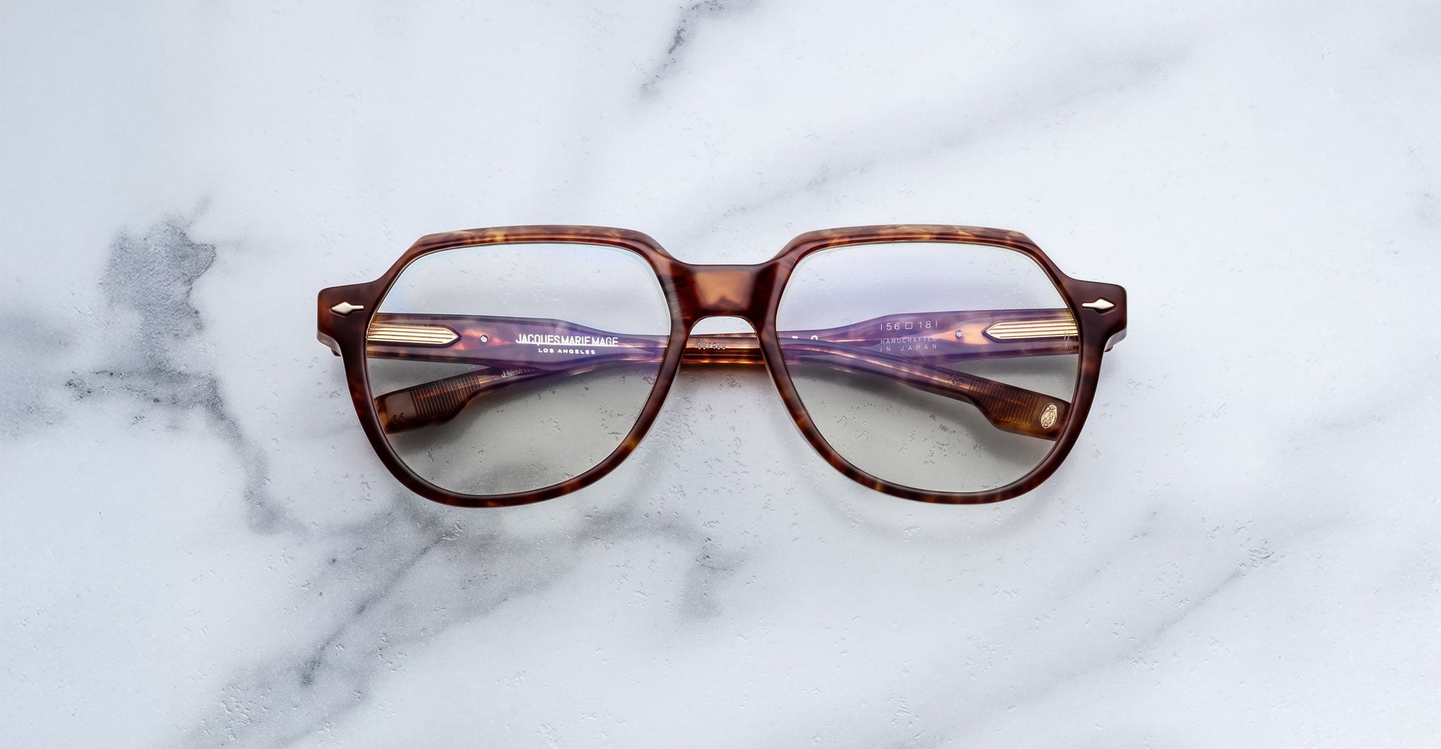 The Jacques Marie Mage Shozo eyeglasses, featuring a brown geometric frame and purple-tinted arms, rest on a white marble surface.