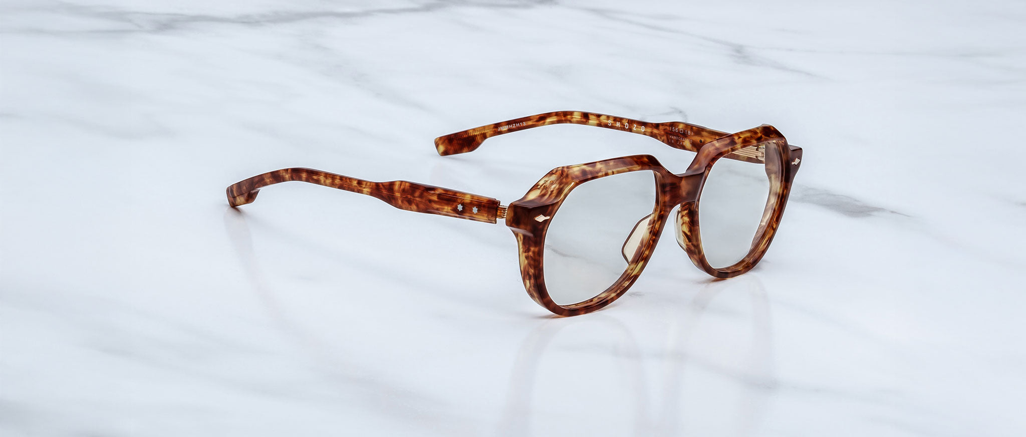 A pair of Jacques Marie Mage Shozo geometric brown tortoiseshell eyeglasses rests on a white marble surface.