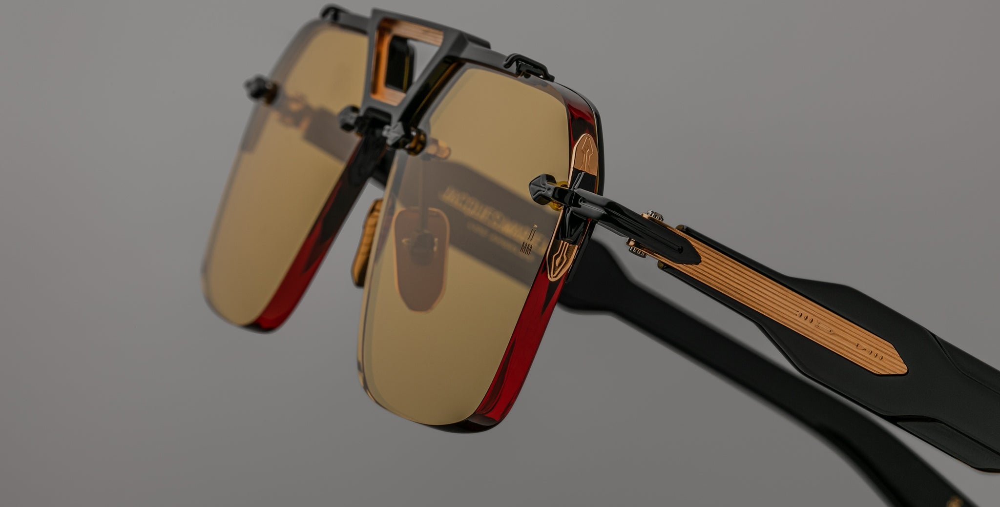 Close-up of Jacques Marie Mage Silverton sunglasses featuring rectangular yellow-tinted lenses, black and red frames, and metallic gold accents on the arms, set against a plain gray background.