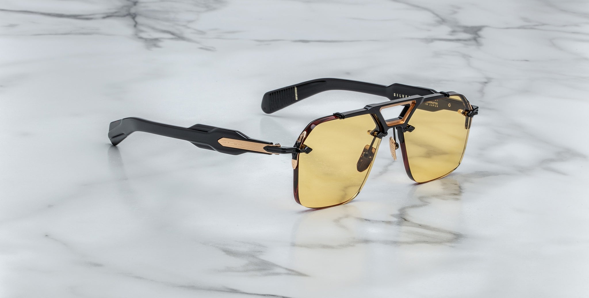 The Jacques Marie Mage Silverton sunglasses feature rectangular yellow-tinted lenses, thick black arms with gold accents, and are displayed on a white marble surface.