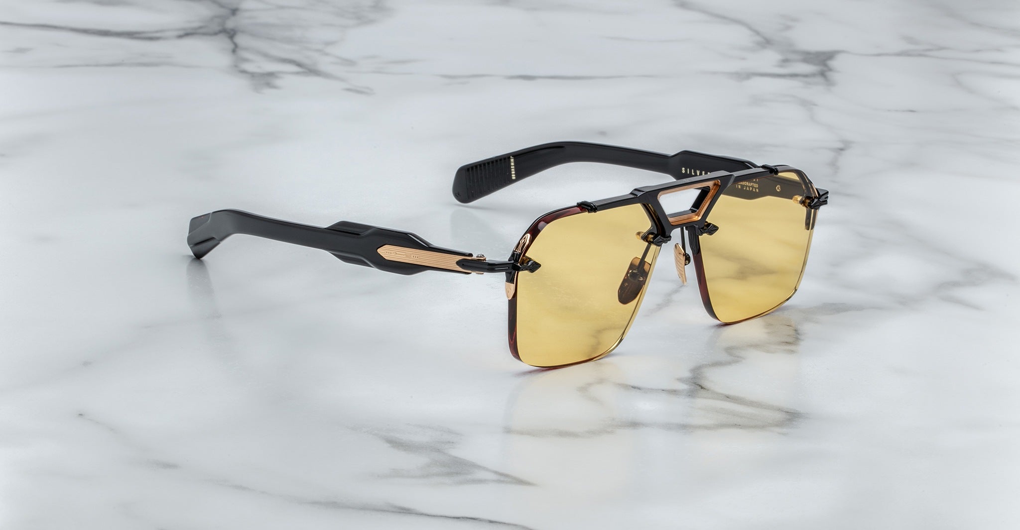 The Jacques Marie Mage Silverton sunglasses feature rectangular yellow-tinted lenses, thick black arms with gold accents, and are displayed on a white marble surface.