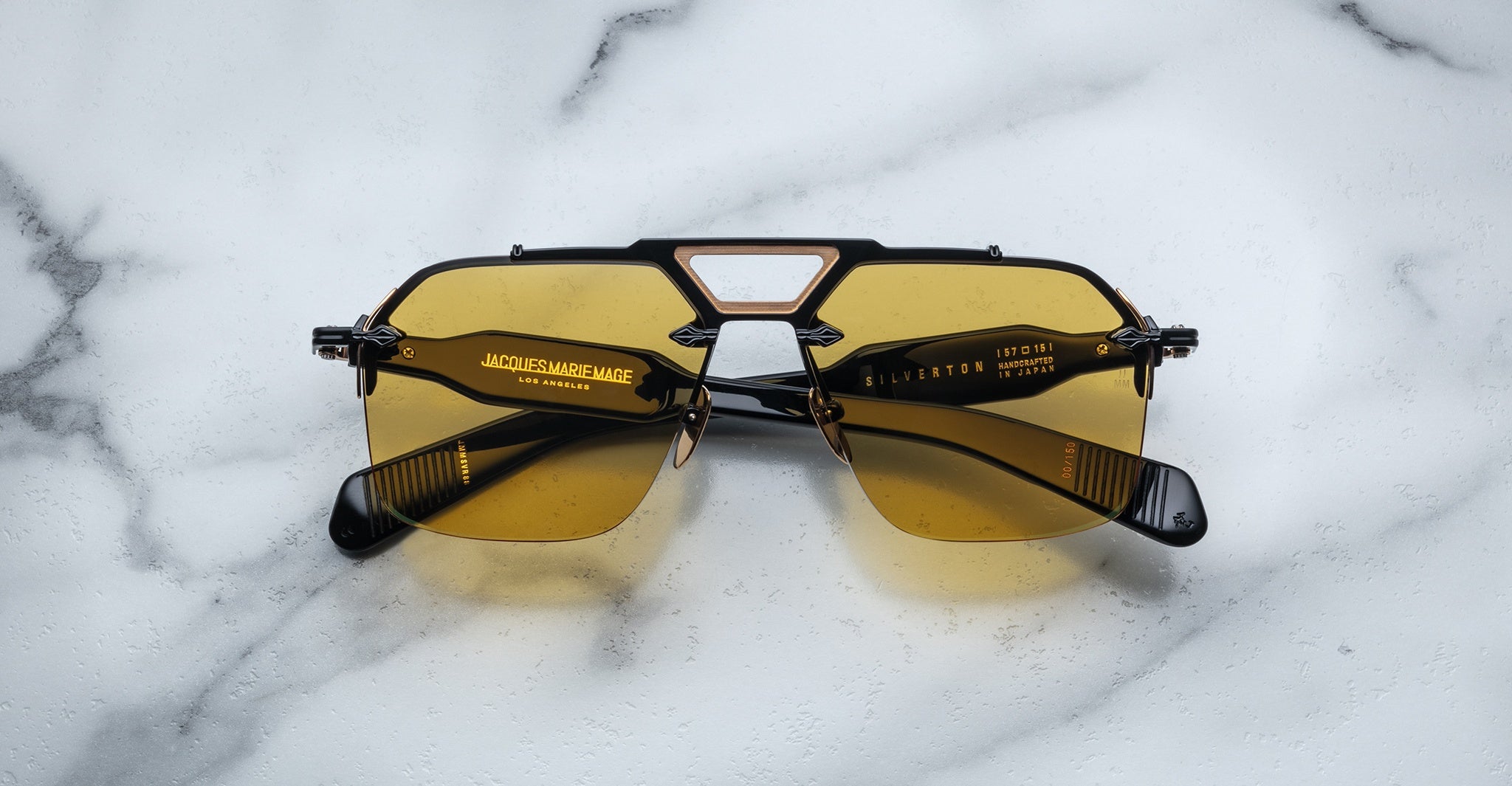 A pair of Jacques Marie Mage Silverton black sunglasses with yellow-tinted lenses rests on a white marble surface. Gold brand markings and text appear on the inner sides of the arms.