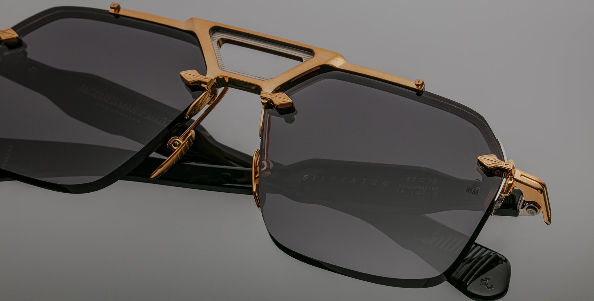 A close-up of Jacques Marie Mage Silverton black sunglasses with dark tinted lenses and gold metallic accents on the frame and bridge, set against a plain gray background.