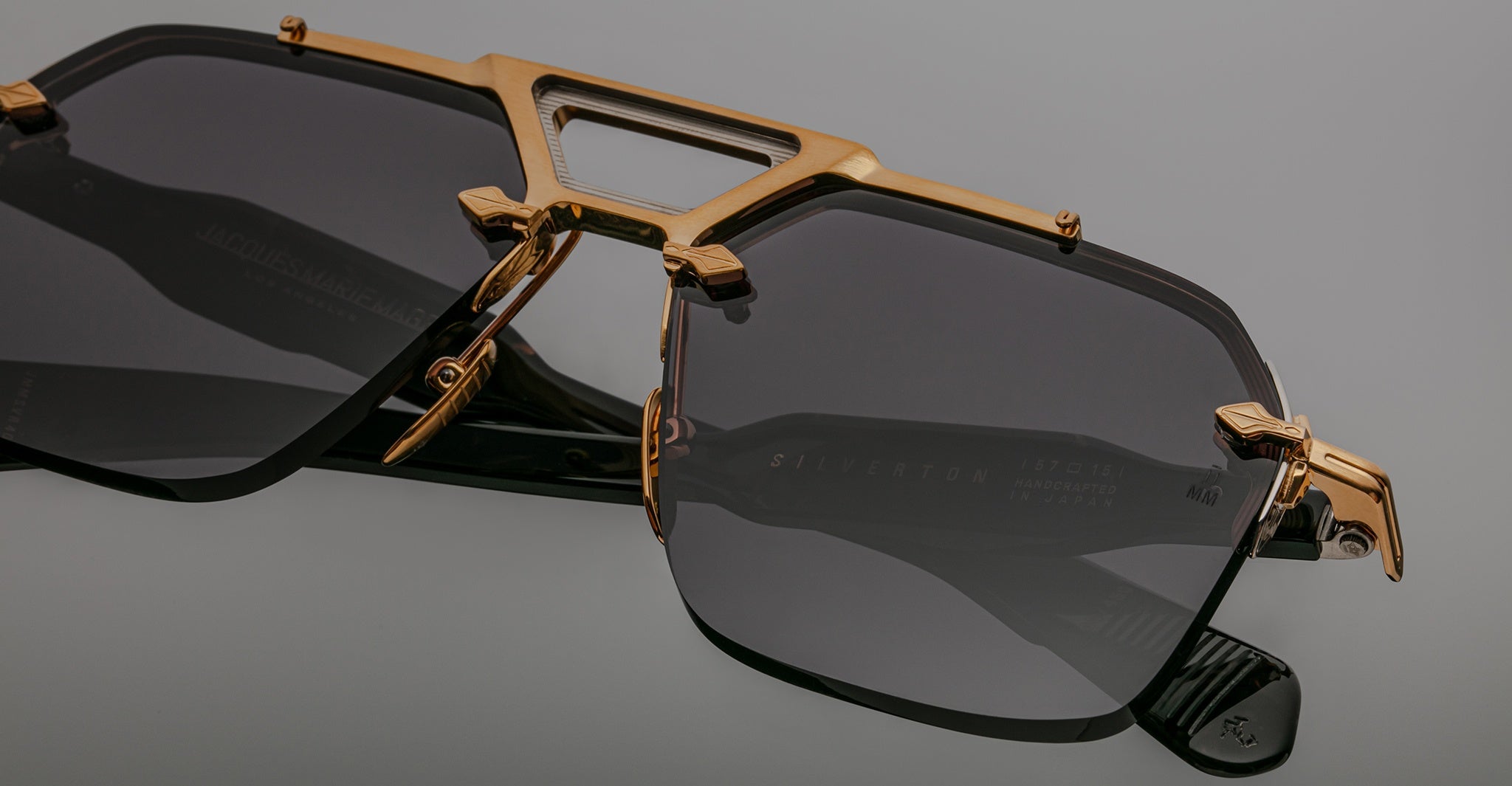A close-up of Jacques Marie Mage Silverton black sunglasses with dark tinted lenses and gold metallic accents on the frame and bridge, set against a plain gray background.