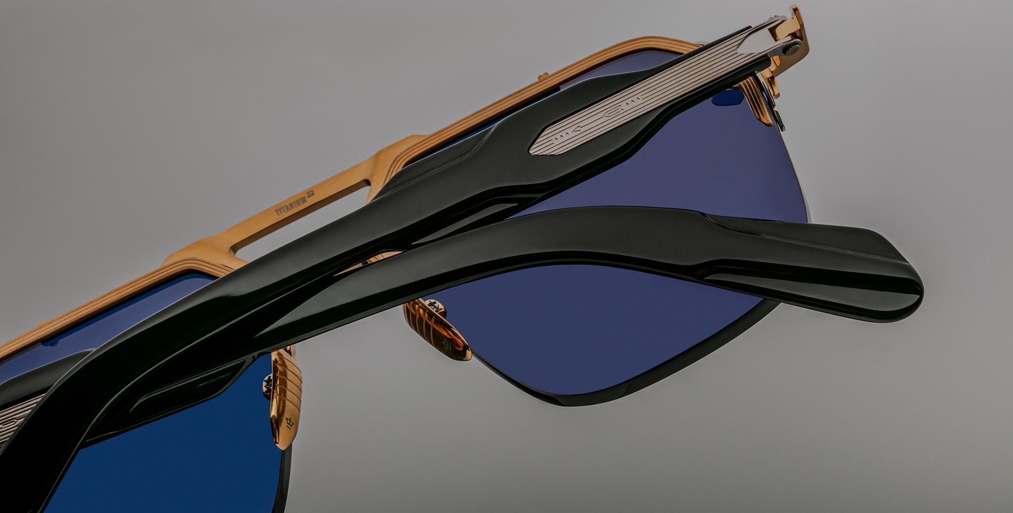 Close-up view of Jacques Marie Mage Silverton sunglasses featuring gold frames, blue-tinted lenses, and black arms, set against a plain gray background.