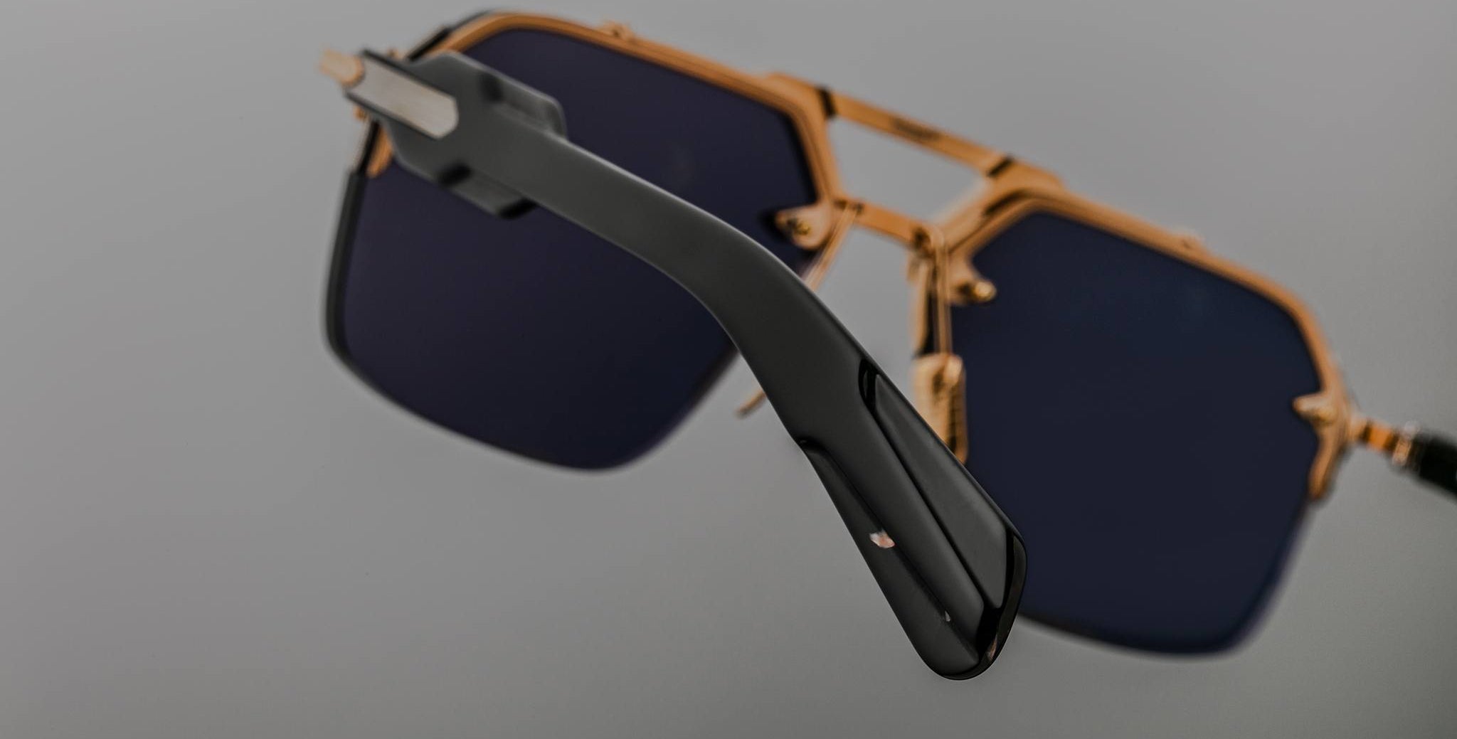 Close-up of Jacques Marie Mage Silverton sunglasses, featuring dark lenses and a gold frame. The focus is on the black temple arm in the foreground, while the lenses and frame appear slightly blurred in the background.