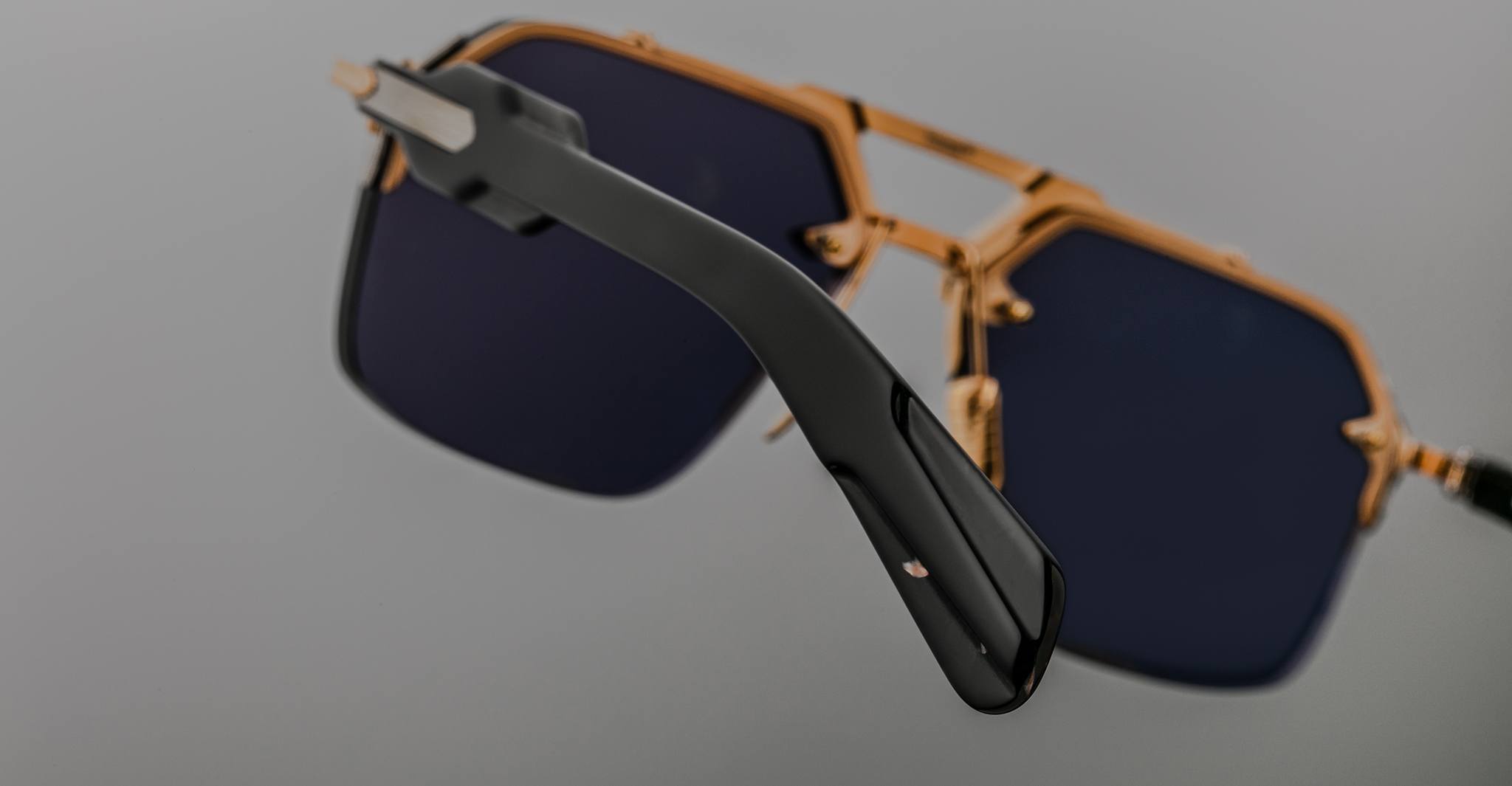 Close-up of Jacques Marie Mage Silverton sunglasses, featuring dark lenses and a gold frame. The focus is on the black temple arm in the foreground, while the lenses and frame appear slightly blurred in the background.